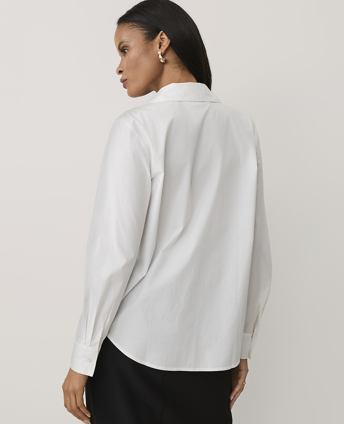 A woman wearing a white long-sleeved button-down shirt and black trousers stands against a plain, light-colored background.