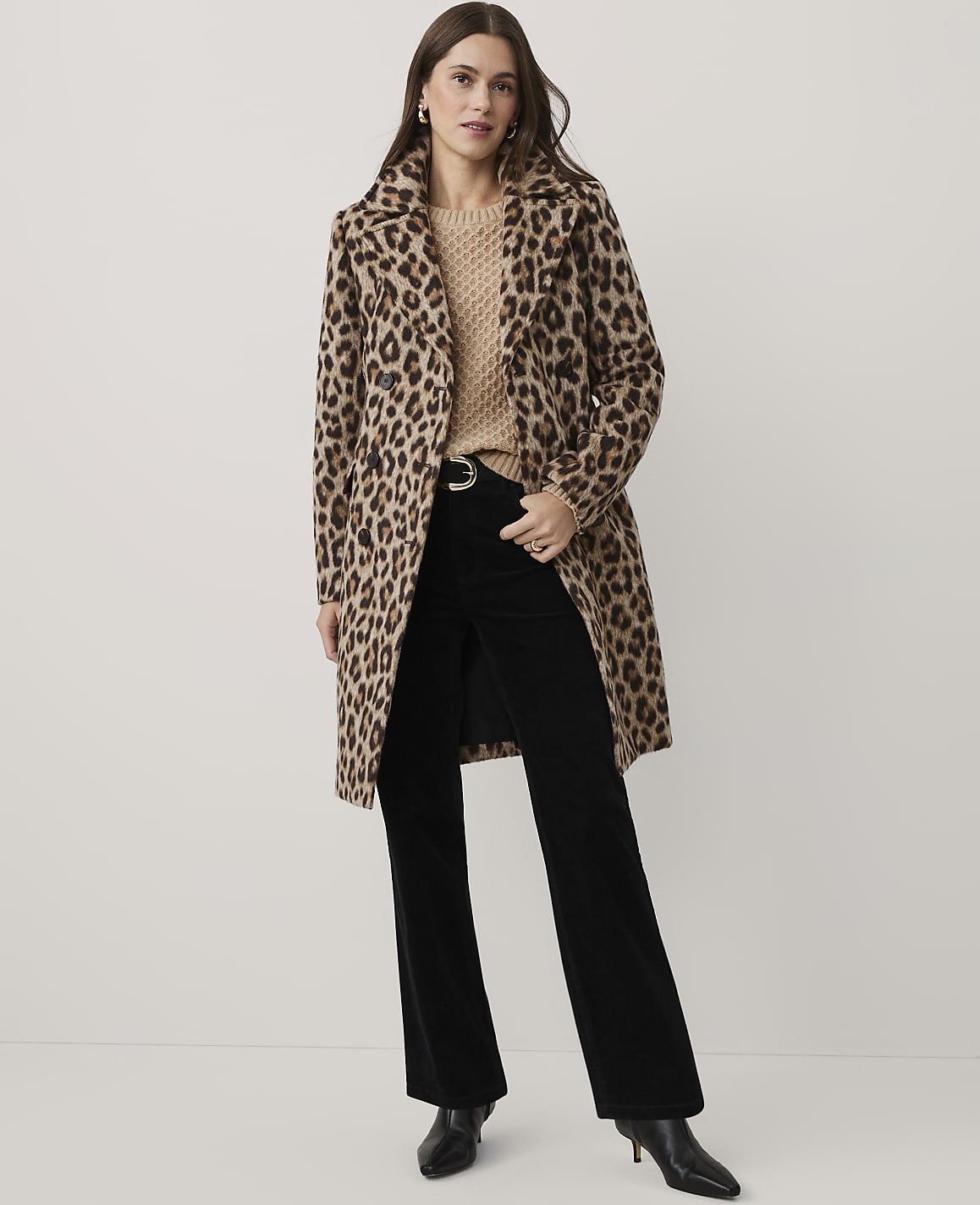 A woman wearing a leopard print coat, beige sweater, and black flared trousers stands against a plain white background.