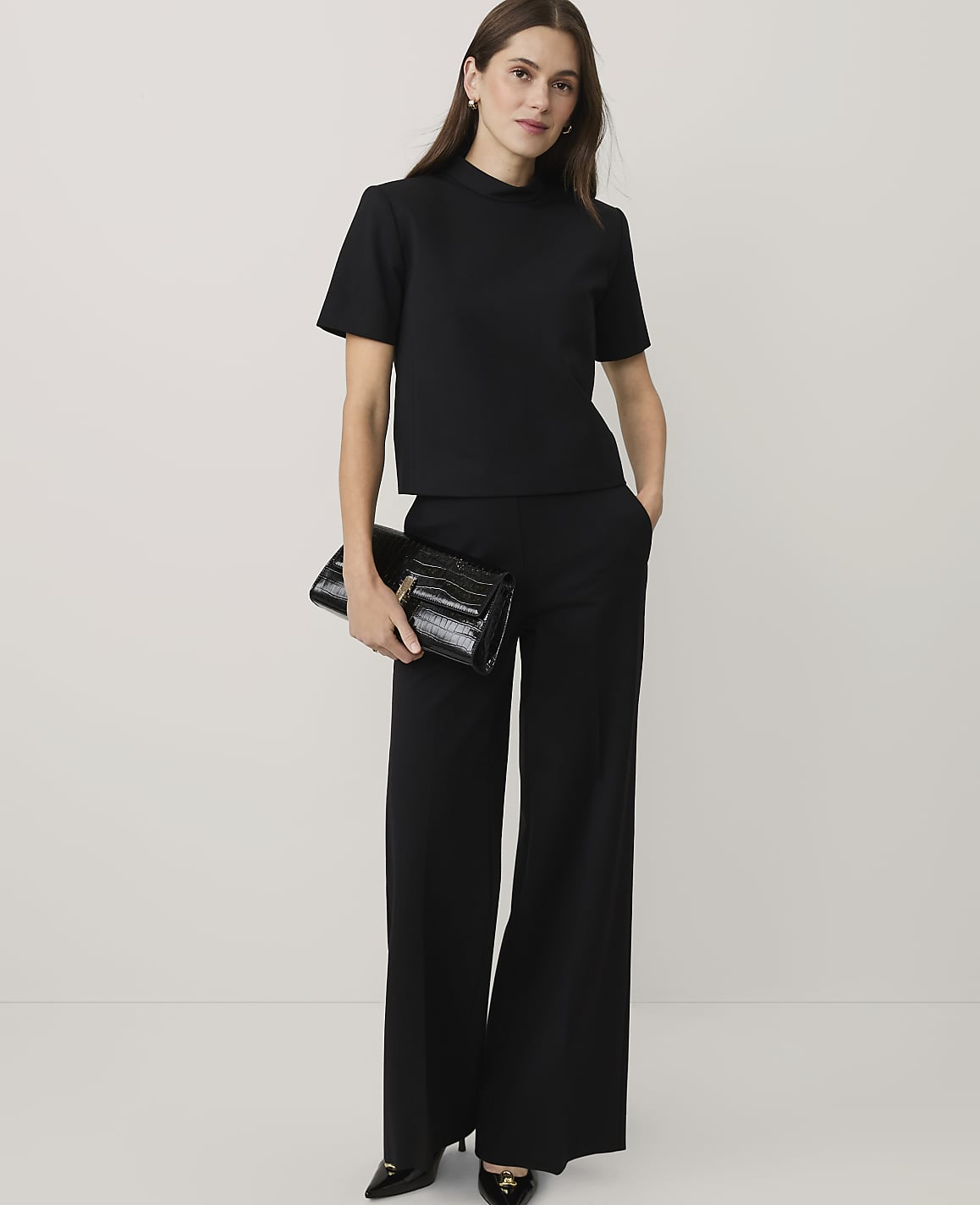 A woman wearing a black short-sleeved top and wide-leg trousers holds a black clutch against a plain white background.