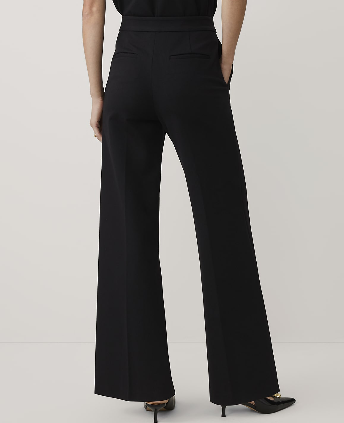 A person wearing wide-leg black trousers and black heels stands against a plain, light-colored background.