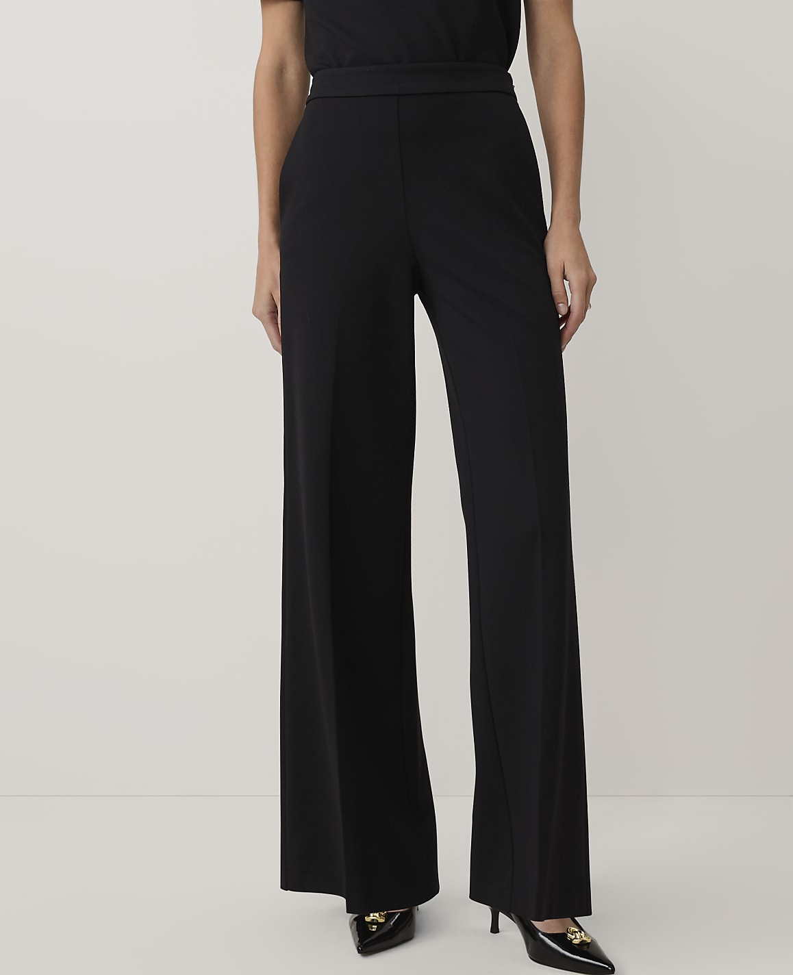 A person wearing black wide-leg trousers and pointed heels stands against a plain white background.