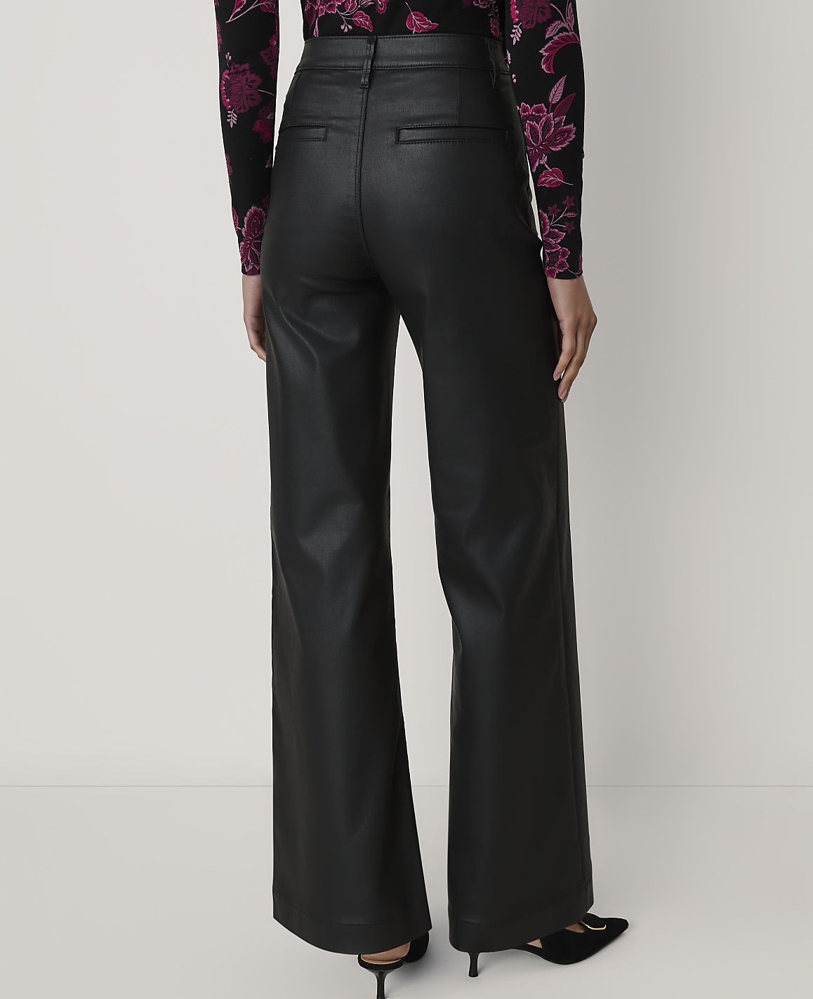 A person wearing black leather wide-leg trousers and a floral top stands against a plain white background.