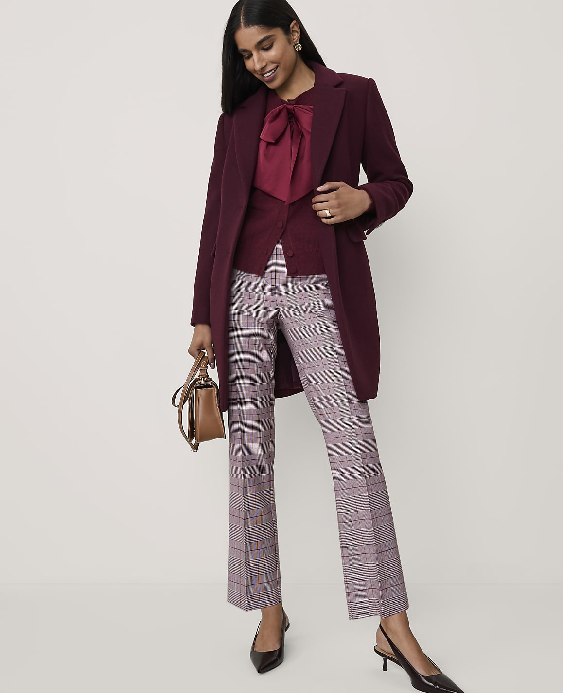 A woman wearing a burgundy coat and plaid trousers stands against a plain light grey background.