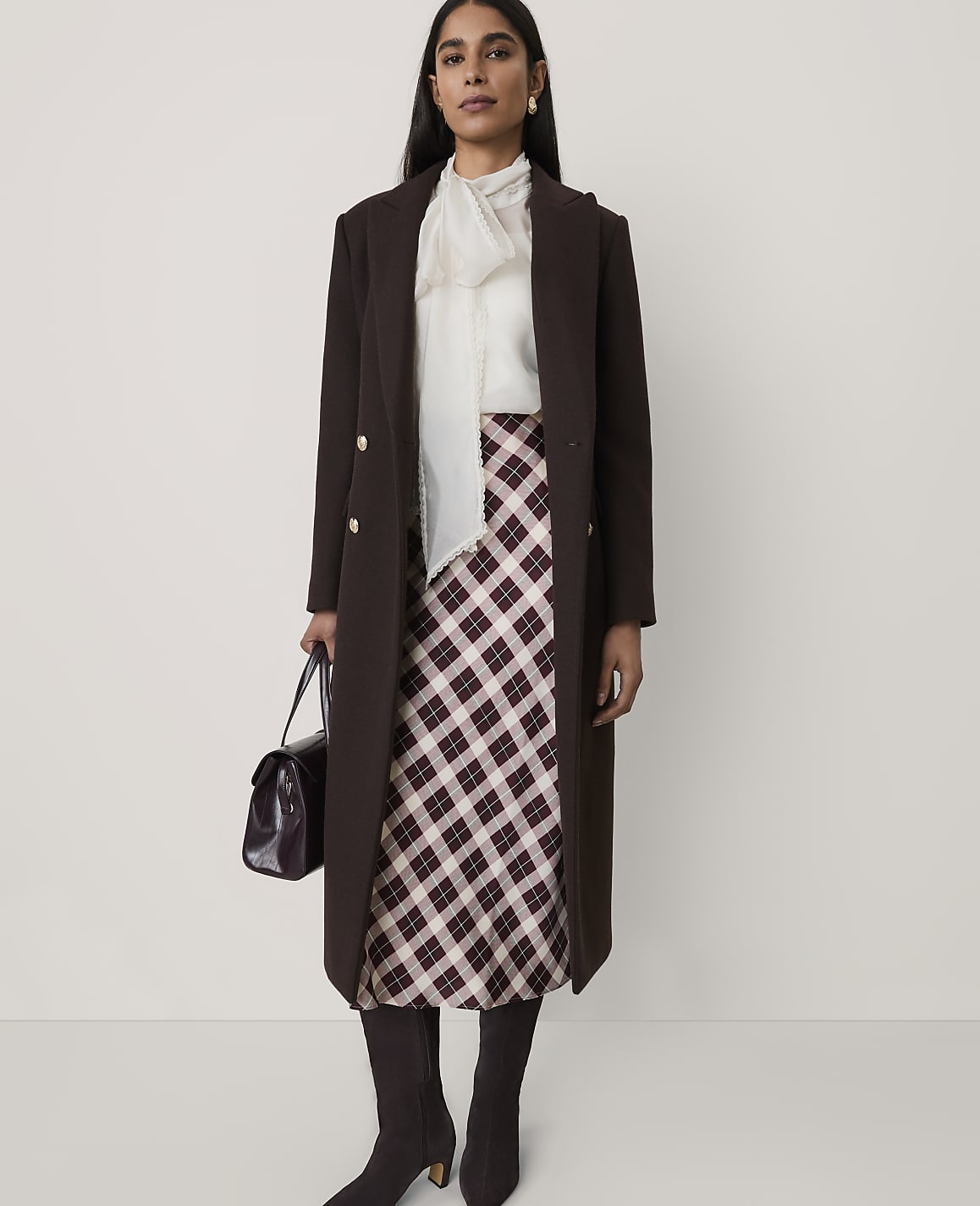 A woman wearing a long brown coat, a white blouse, and a plaid skirt stands against a plain, light-colored background.
