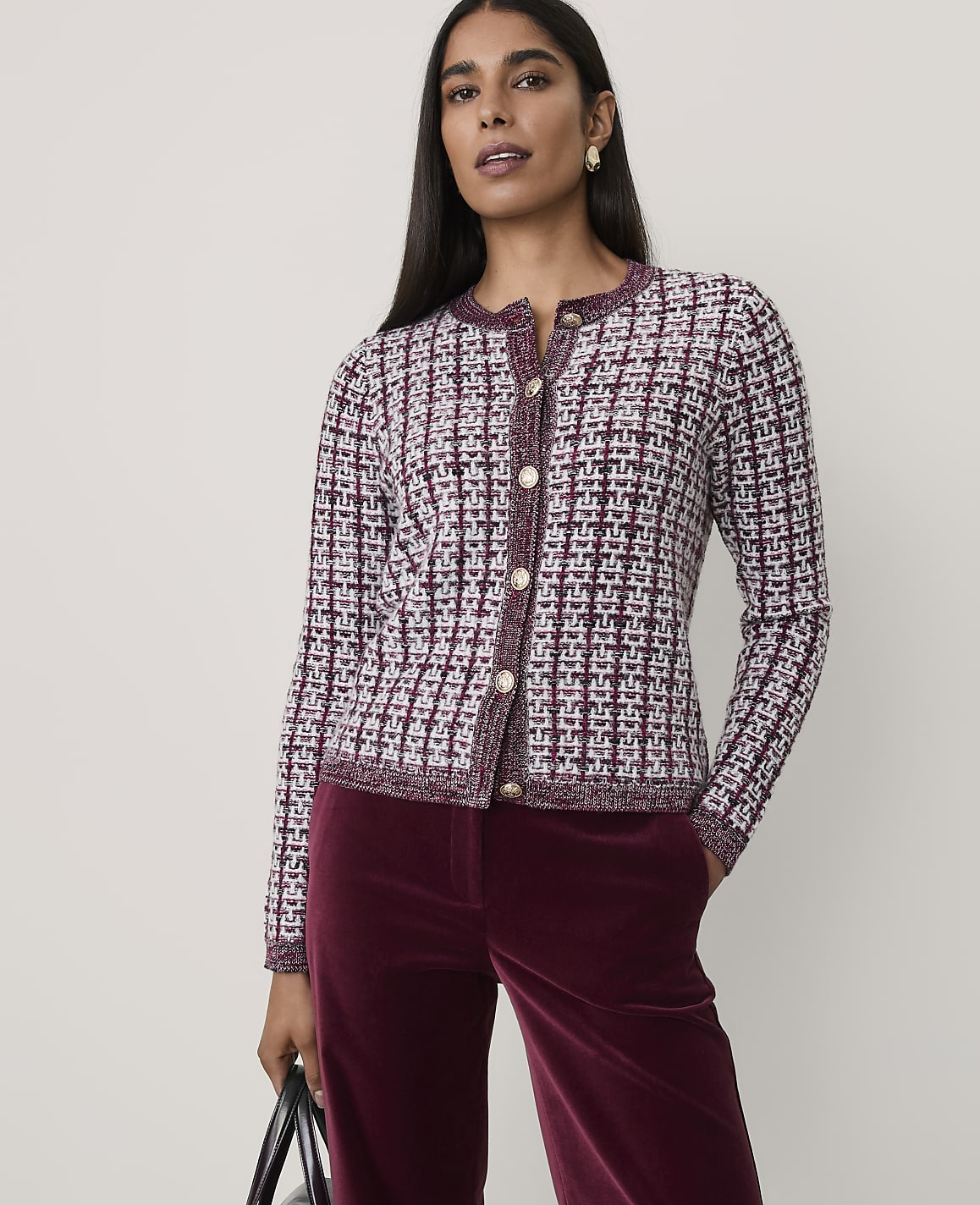 A woman wearing a purple and white tweed jacket and burgundy velvet trousers stands against a plain, light-colored background.