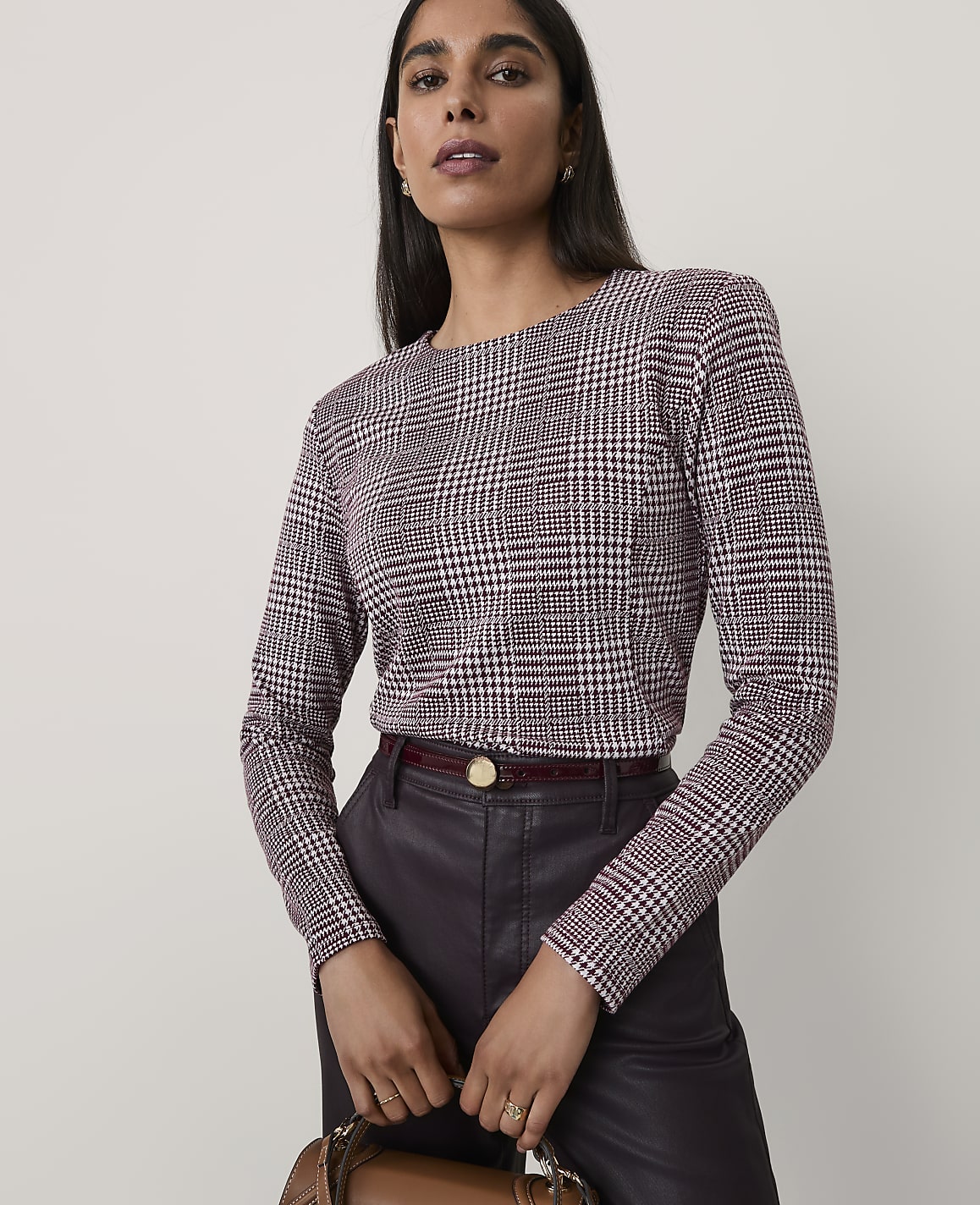 A woman wearing a maroon and white houndstooth top and dark leather trousers stands against a plain, light-colored background.