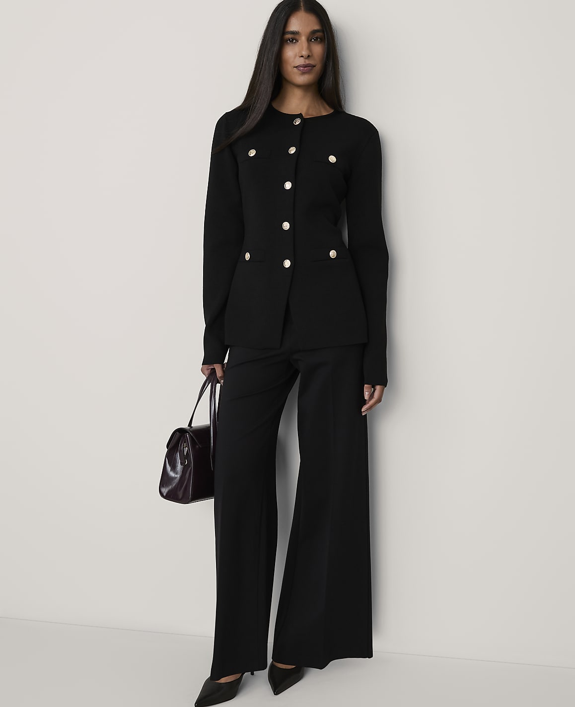 A woman wearing a black buttoned blazer and wide-leg trousers holds a burgundy handbag against a plain white background.
