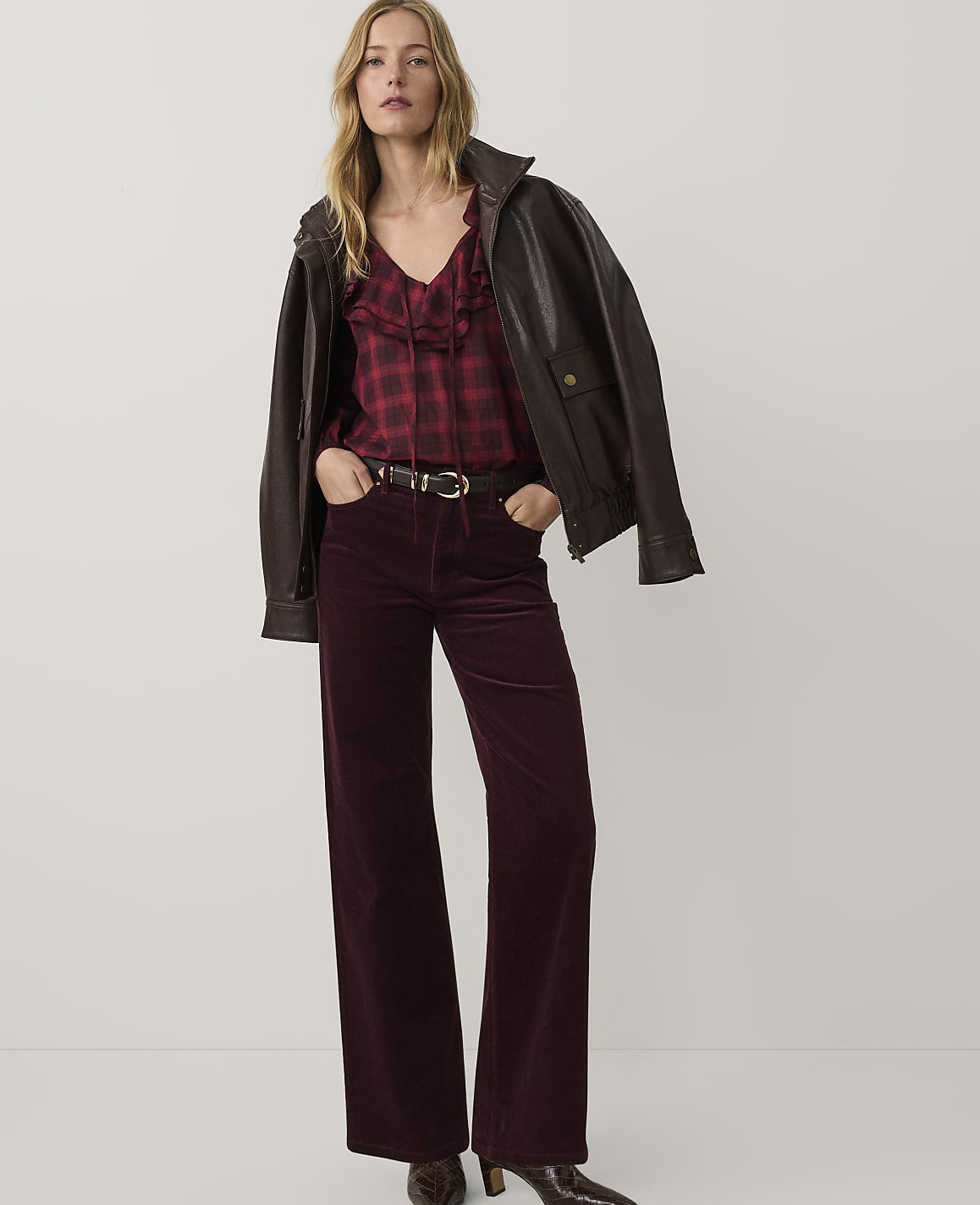 A woman wearing a brown leather jacket, a red plaid shirt, and dark purple flared trousers stands against a plain white background.