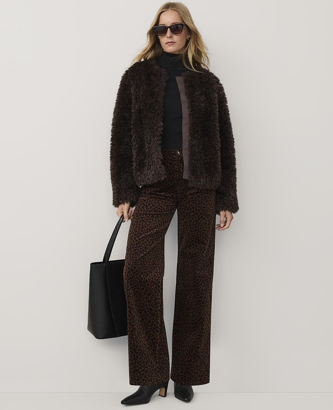 A woman wearing a brown faux-fur jacket, leopard print trousers, and sunglasses stands against a plain white background.