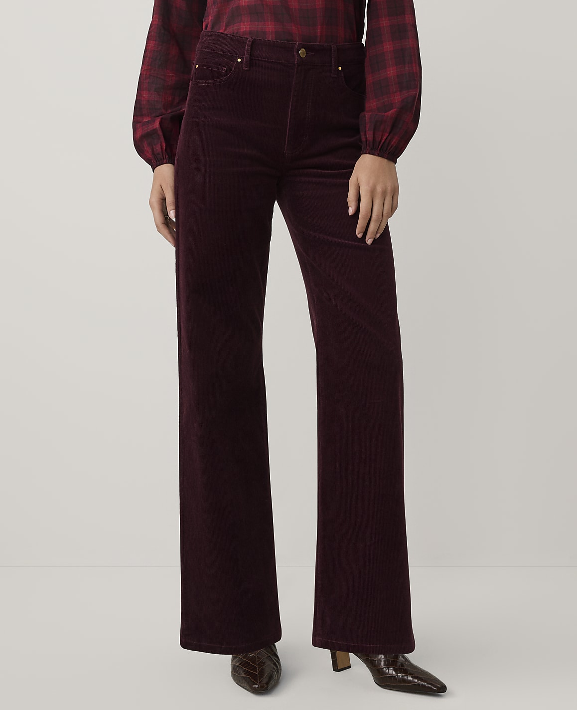 A person wearing a red plaid shirt and burgundy corduroy wide-leg trousers stands against a plain light gray background.