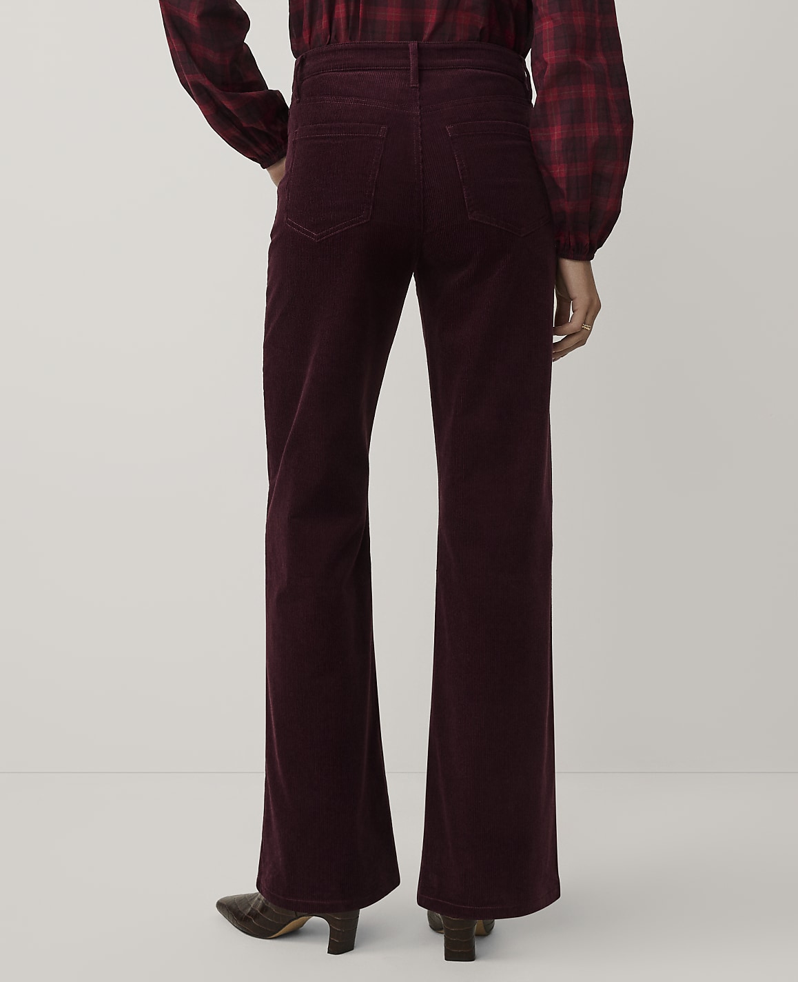 A person wearing a red plaid shirt and burgundy corduroy flare pants stands against a plain white background.