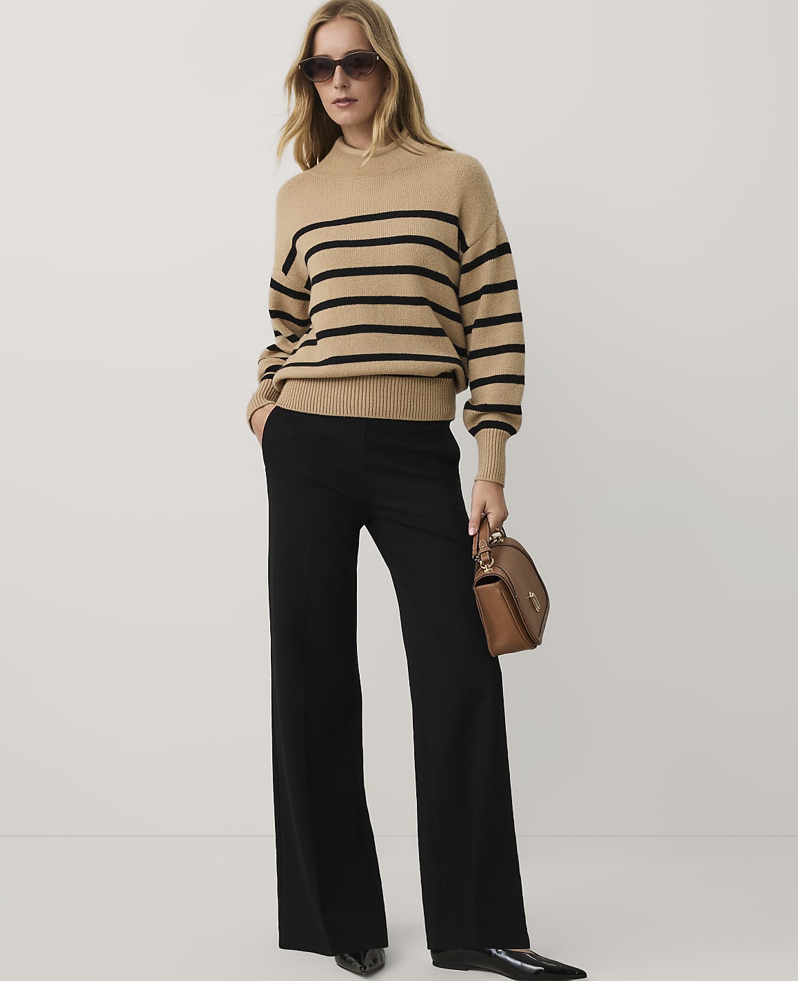A woman wearing a tan striped sweater, black wide-leg trousers, and sunglasses stands against a plain white background.