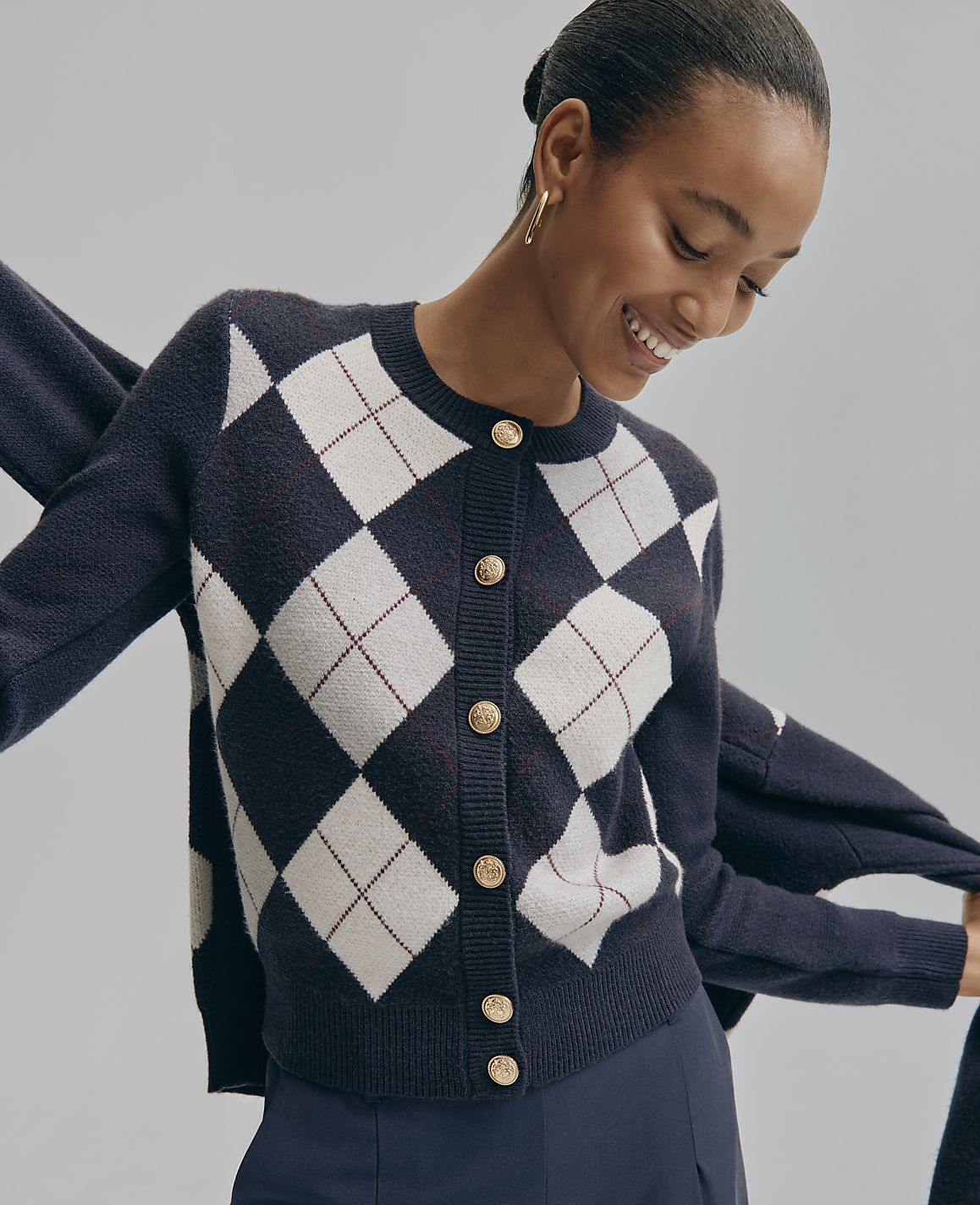 A smiling woman wears a navy and white argyle cardigan against a plain light gray background.