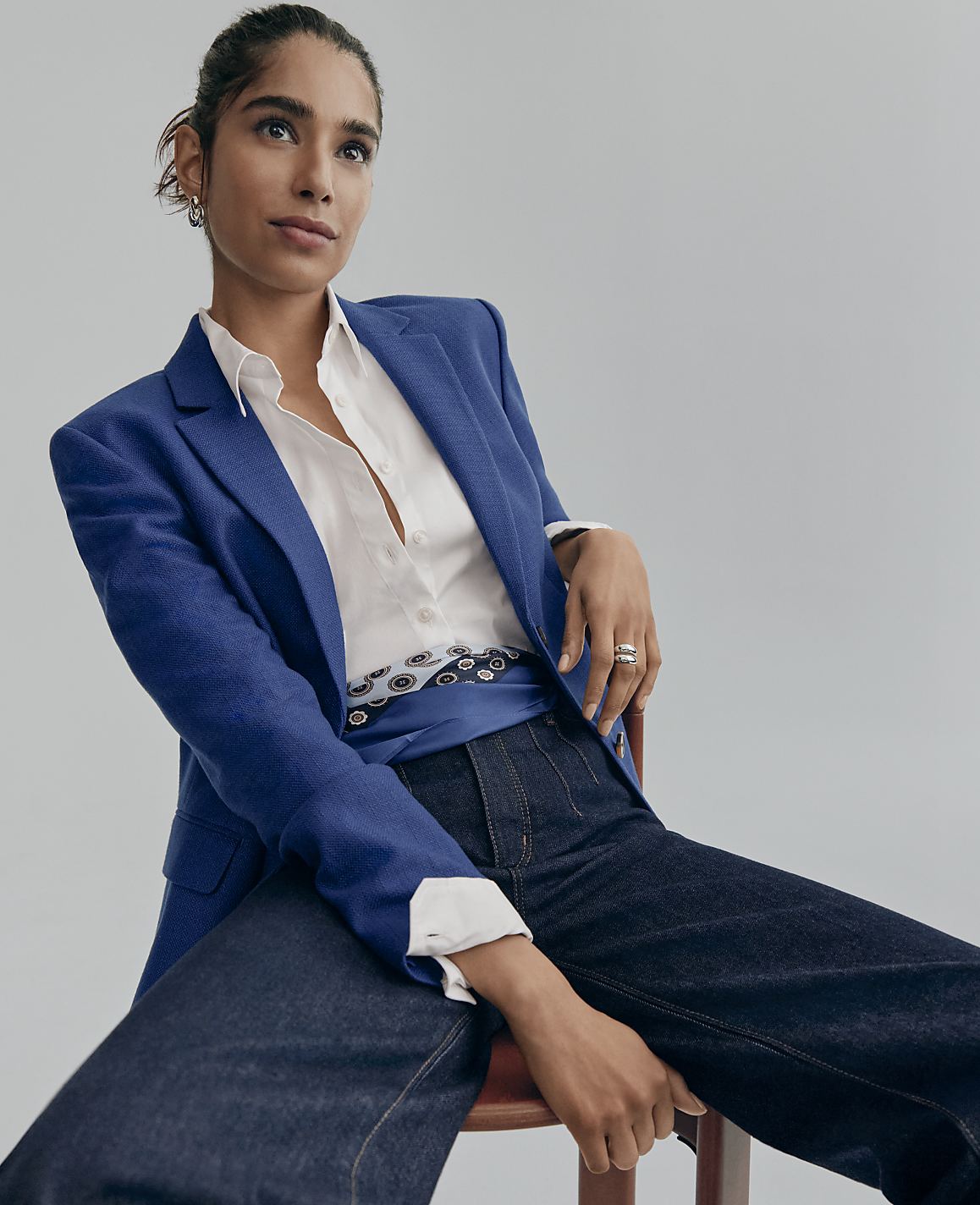 A woman wearing a blue blazer, white shirt, and dark jeans sits on a wooden stool against a plain light gray background.