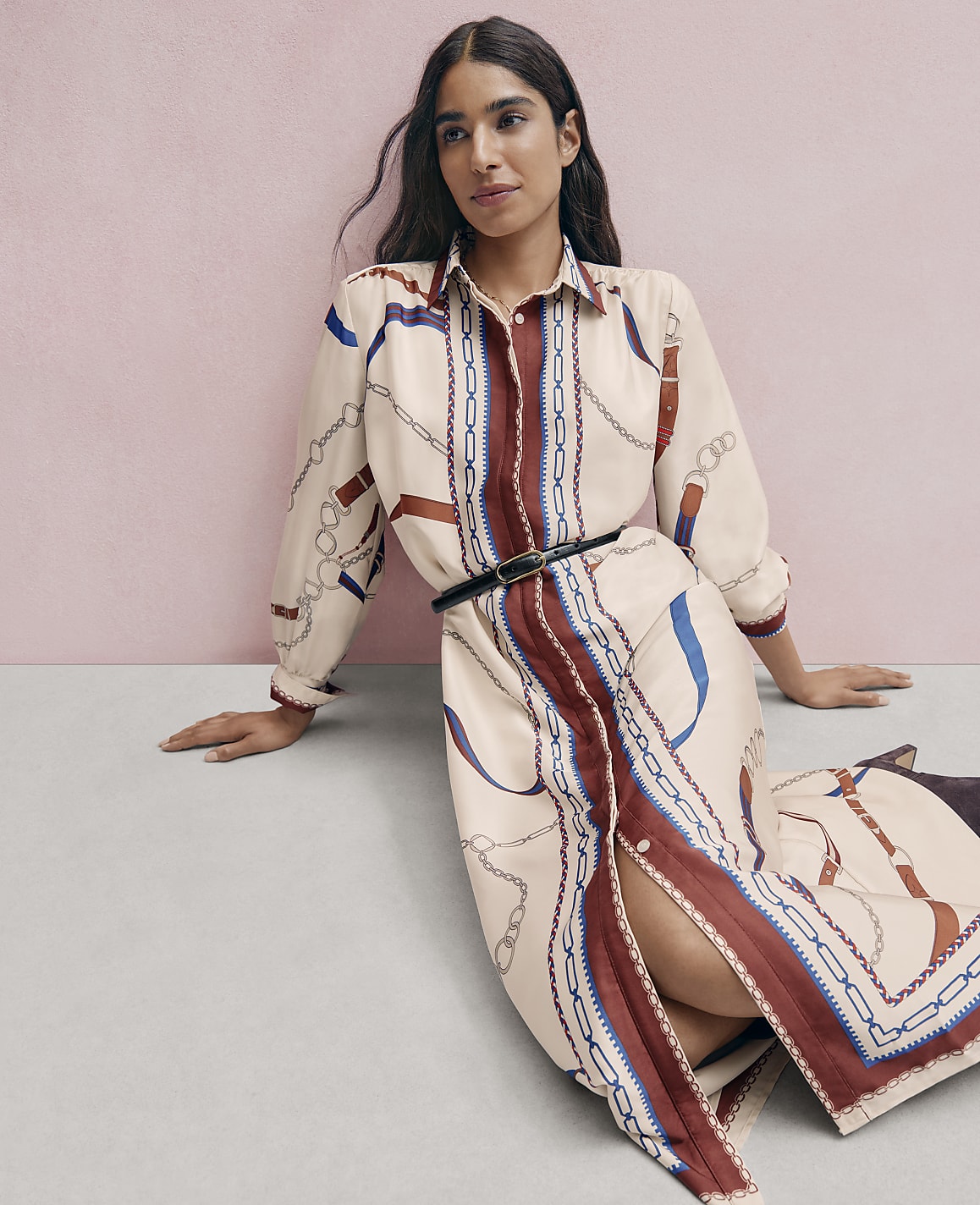 A woman wearing a patterned beige dress and a black belt sits on a grey floor against a pink wall.