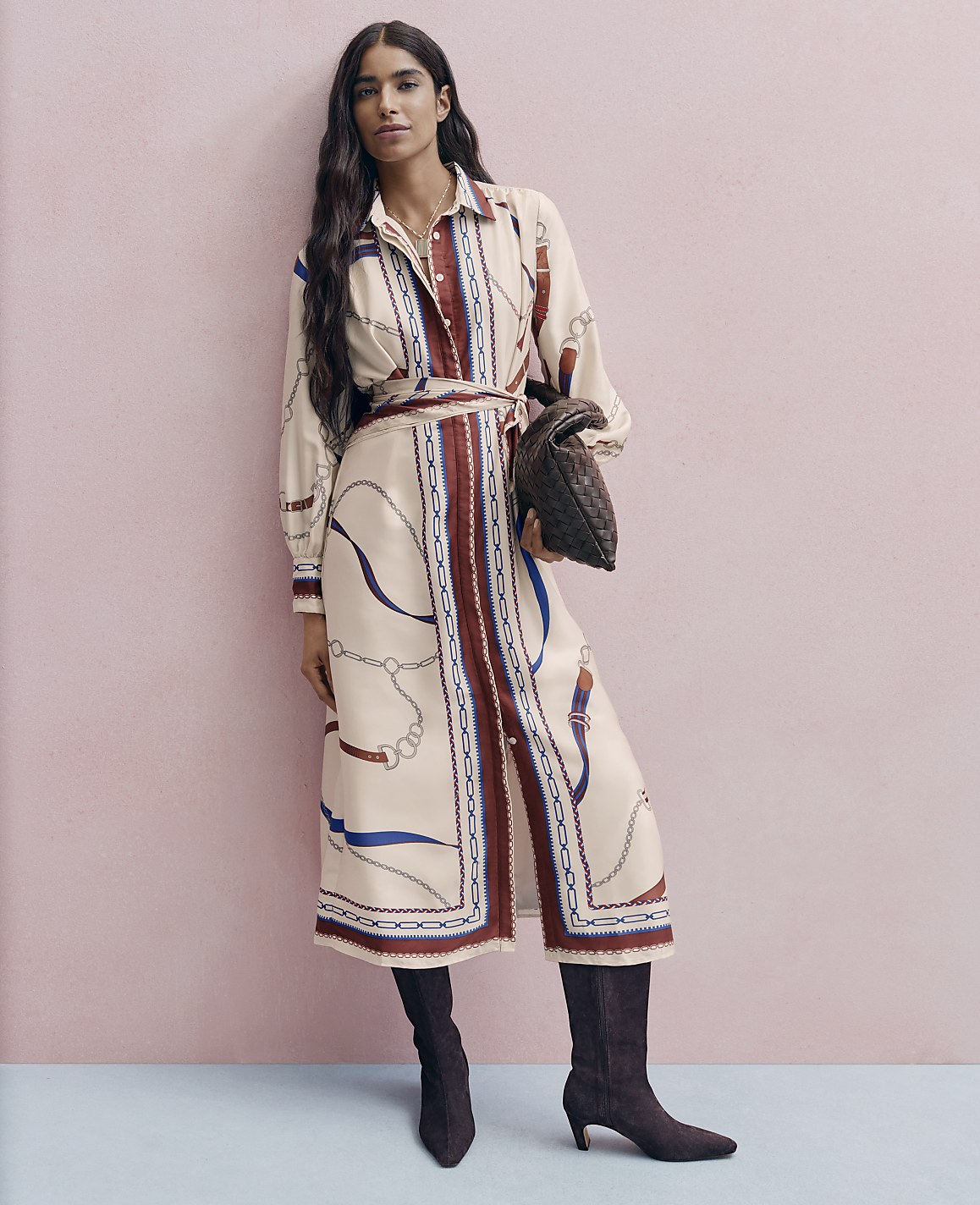 A woman wearing a patterned cream dress and dark brown boots stands against a pink wall and light blue floor.