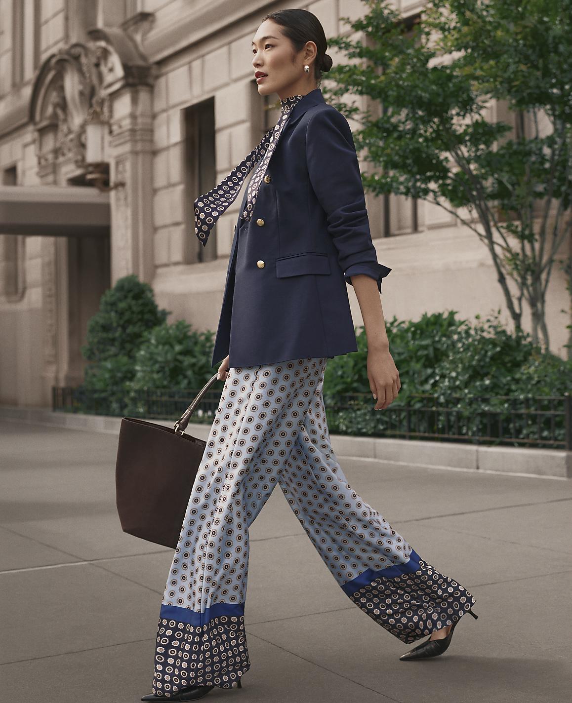 A woman in a navy blazer and patterned wide-leg trousers walks along a city sidewalk in front of a classical stone building and green foliage.