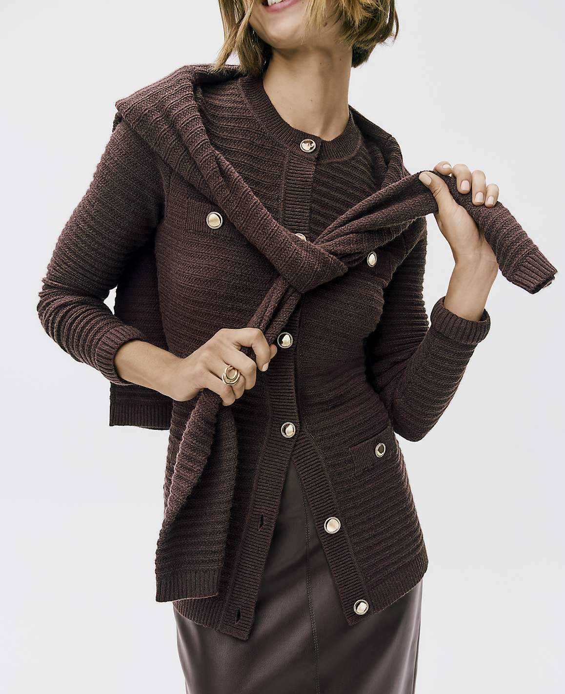 A woman wearing a dark brown ribbed cardigan and leather skirt holds a matching sweater against a plain white background.