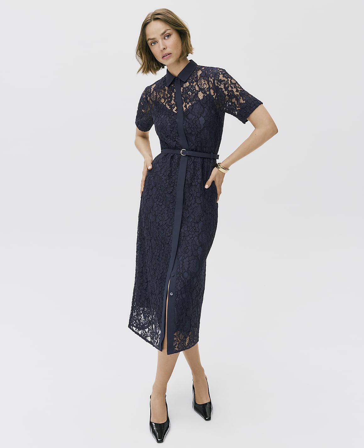A woman wearing a navy blue lace midi dress and black heels stands against a plain white background.