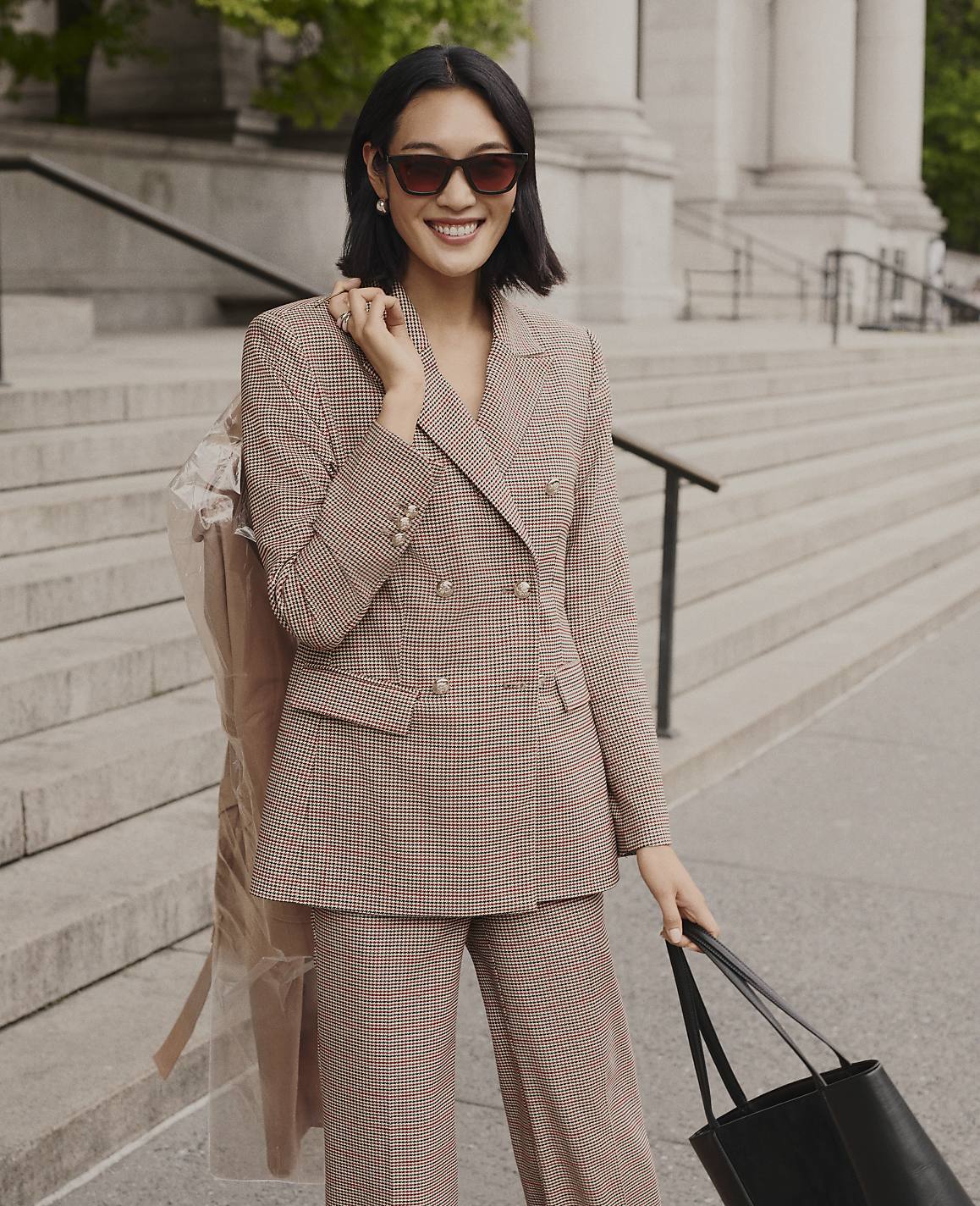 A smiling woman in a brown checkered suit stands on stone steps in front of a building with large white columns.