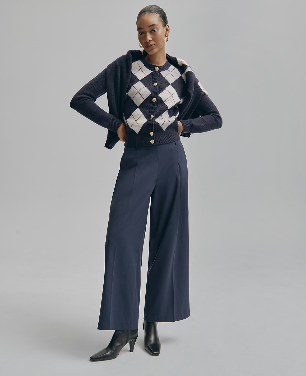 A woman wearing a diamond-patterned cardigan, a dark blue jacket, and wide-leg trousers stands against a plain grey background.