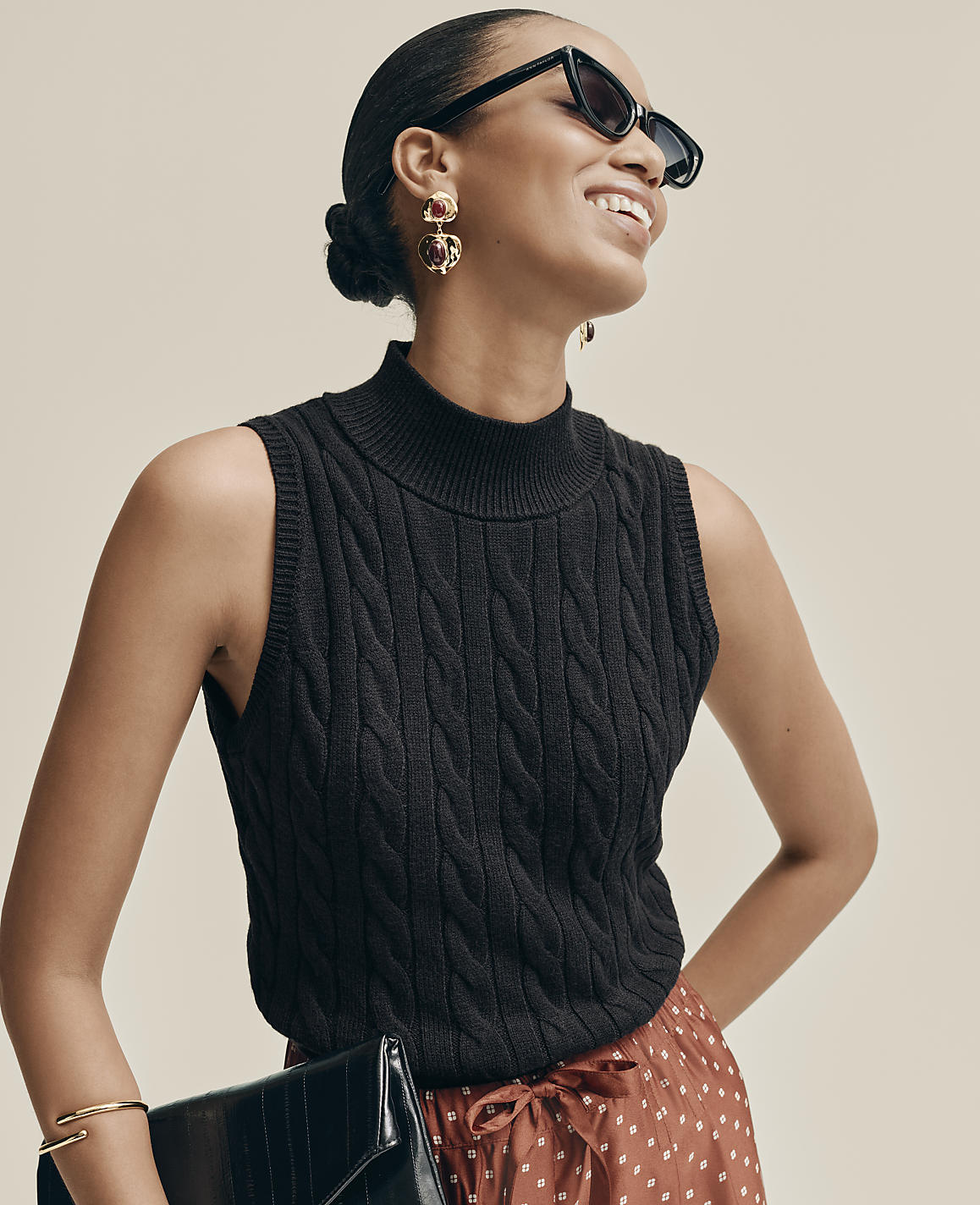 A smiling woman wearing a black cable-knit sleeveless top, sunglasses, and a patterned skirt stands against a plain, neutral background.