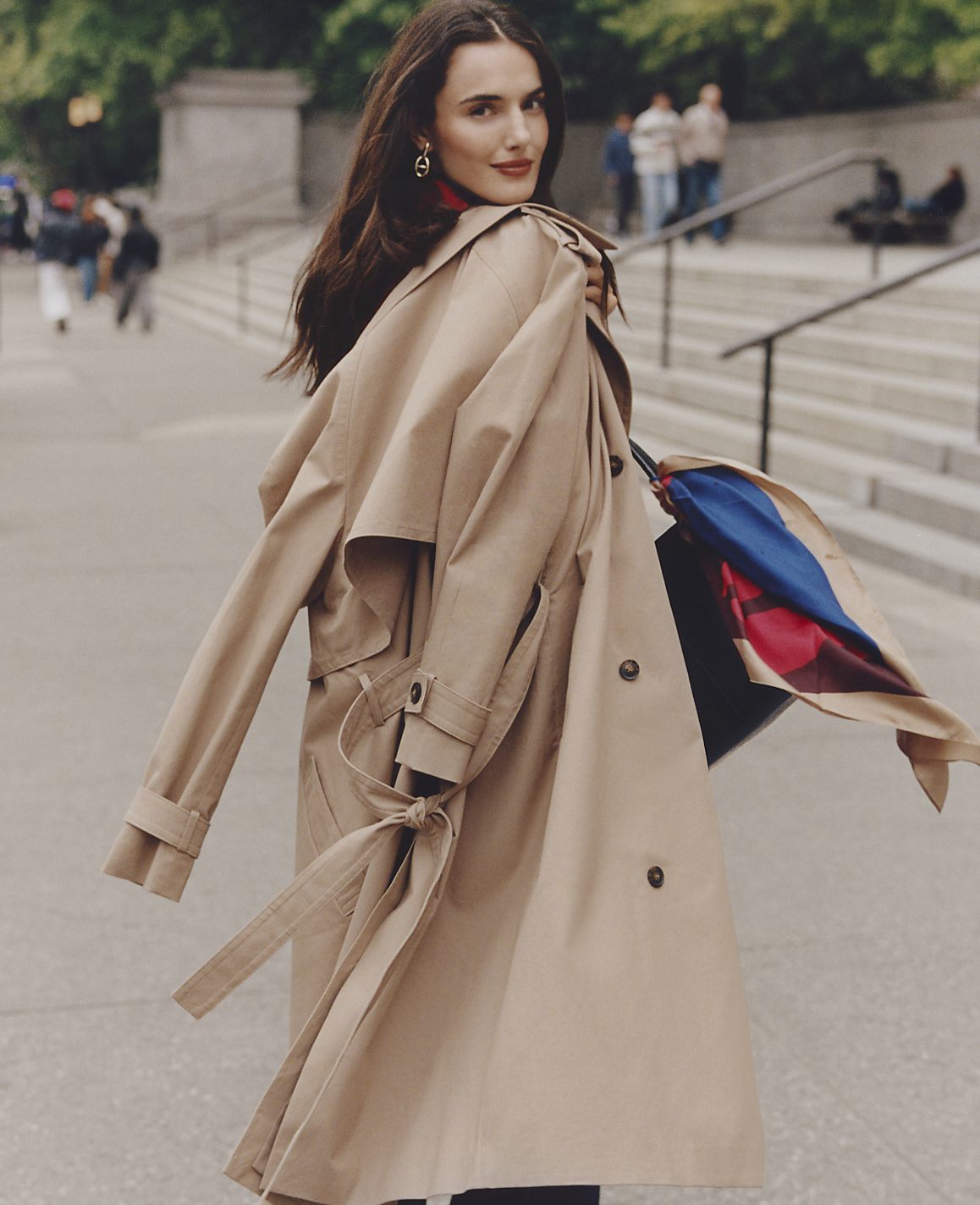 A woman in a tan trench coat stands in the foreground against a blurred city background with stone steps and pedestrians.