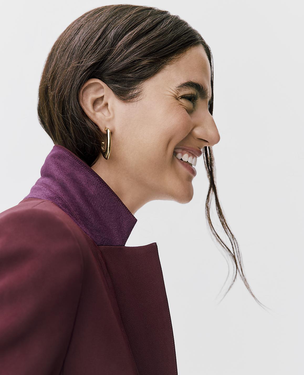 A smiling woman with brown hair and gold hoop earrings wearing a maroon blazer stands against a plain white background.