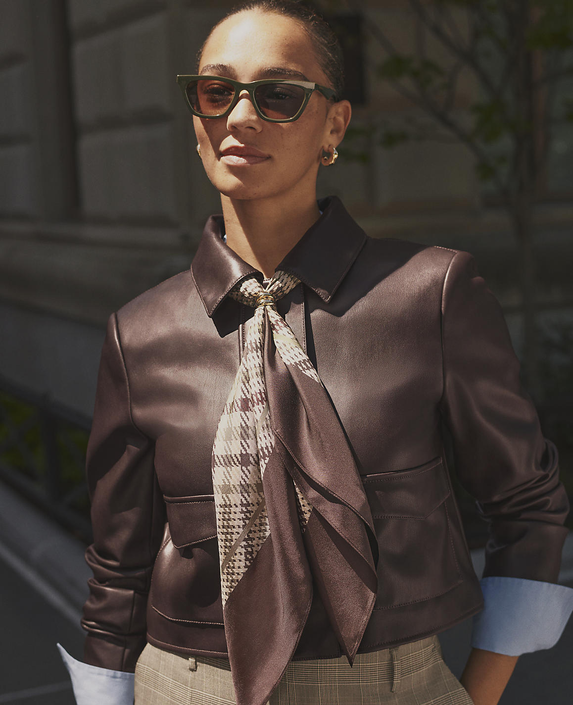 A woman wearing sunglasses, a brown leather jacket, and a patterned scarf stands before a blurred urban building background.