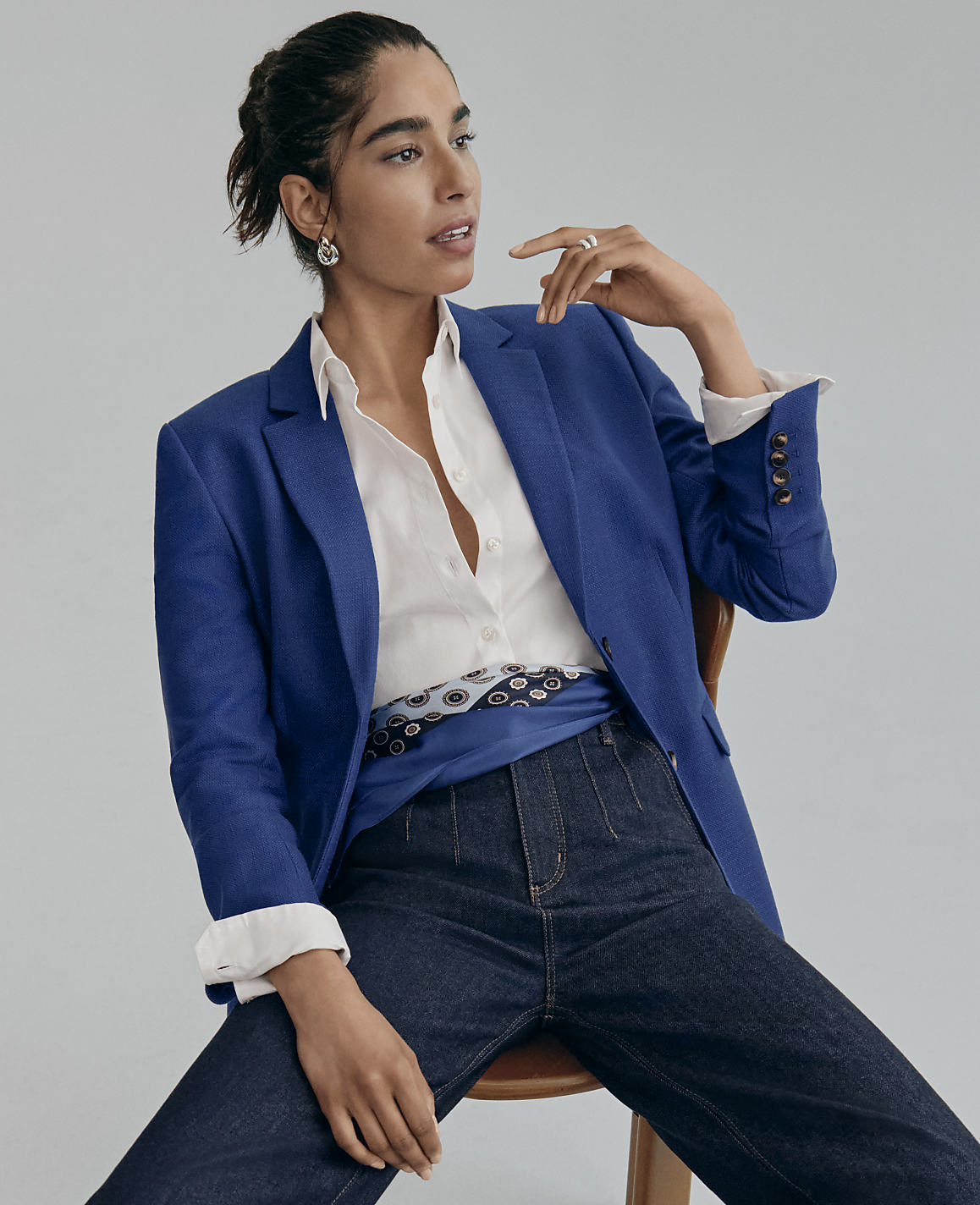 A woman wearing a blue blazer, white shirt, and dark jeans sits on a wooden chair against a plain grey background.