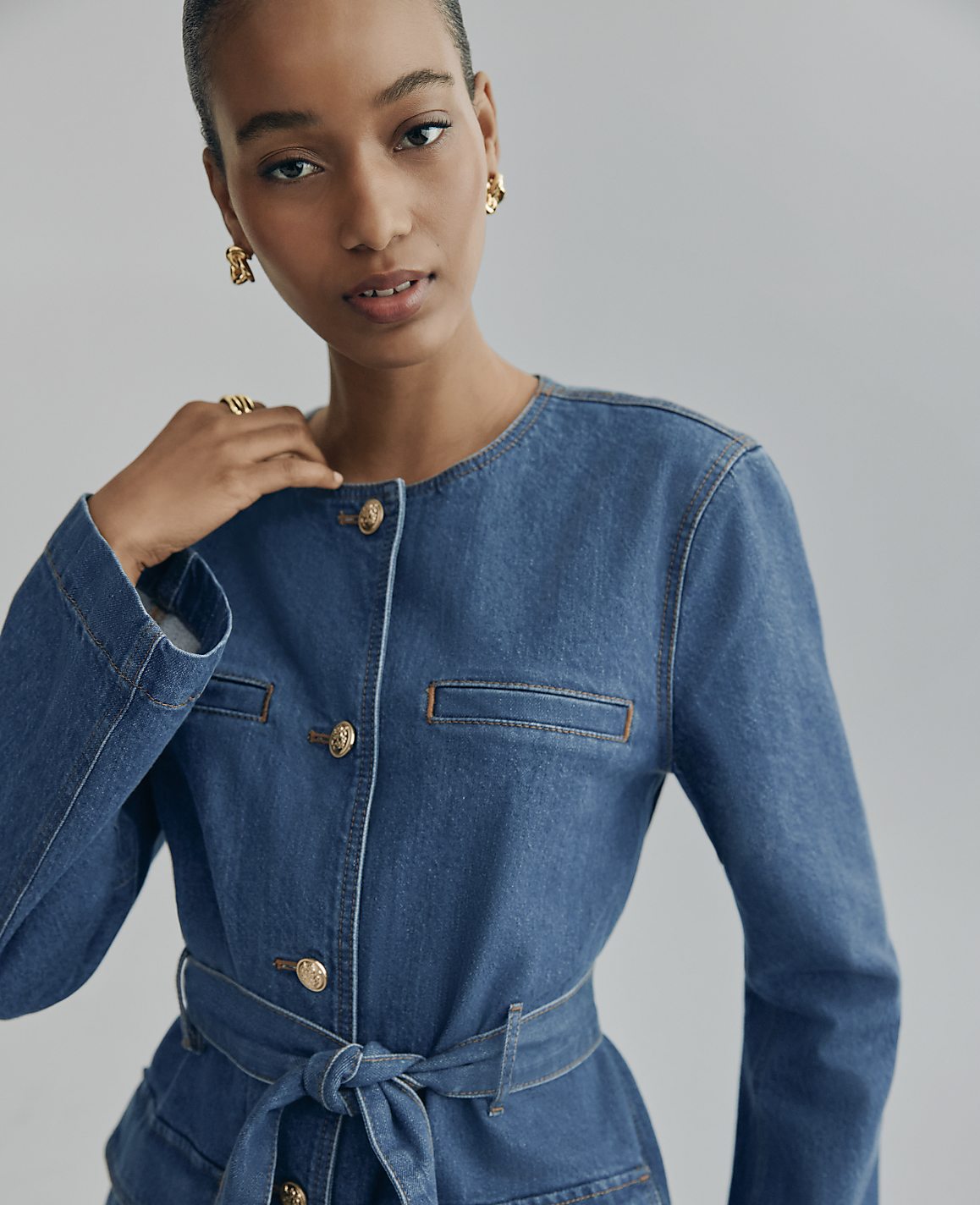 A woman wearing a blue denim jacket and gold earrings poses against a plain grey background.