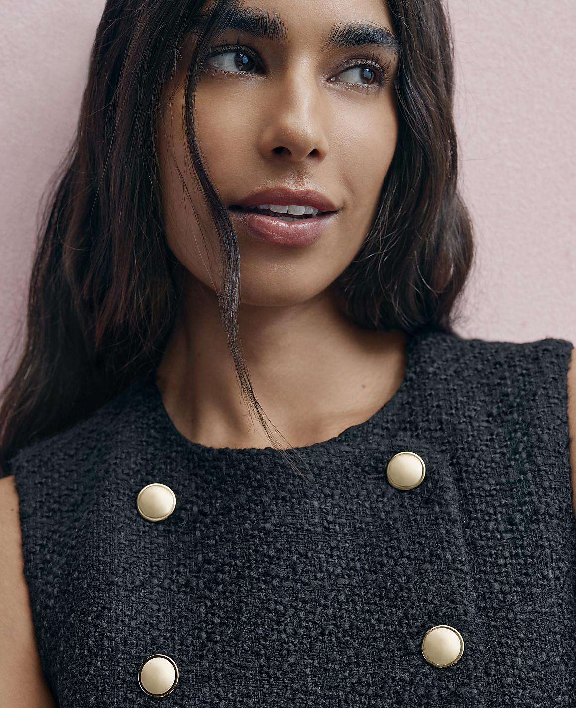 A woman with long dark hair wearing a black textured jacket with gold buttons poses against a light pink wall.
