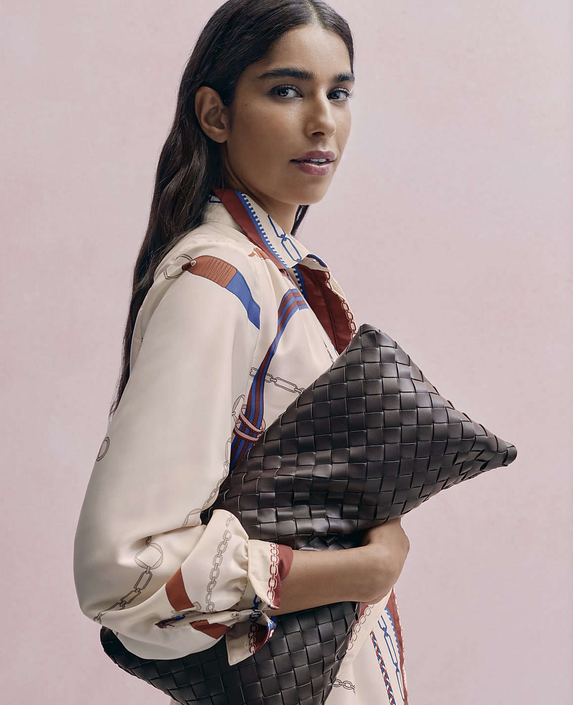 A woman in a patterned cream blouse holds a large woven brown leather clutch against a solid pink background.