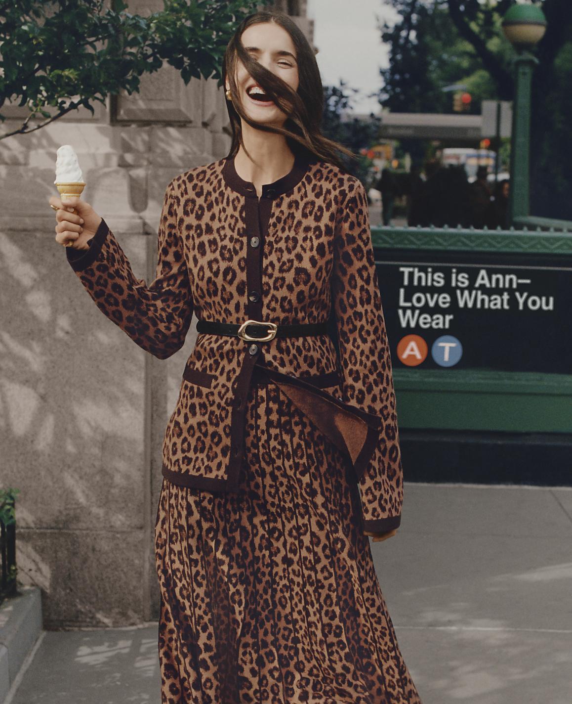 A laughing woman in a leopard-print dress holds an ice cream cone on a city sidewalk with a green sign and trees in the background.