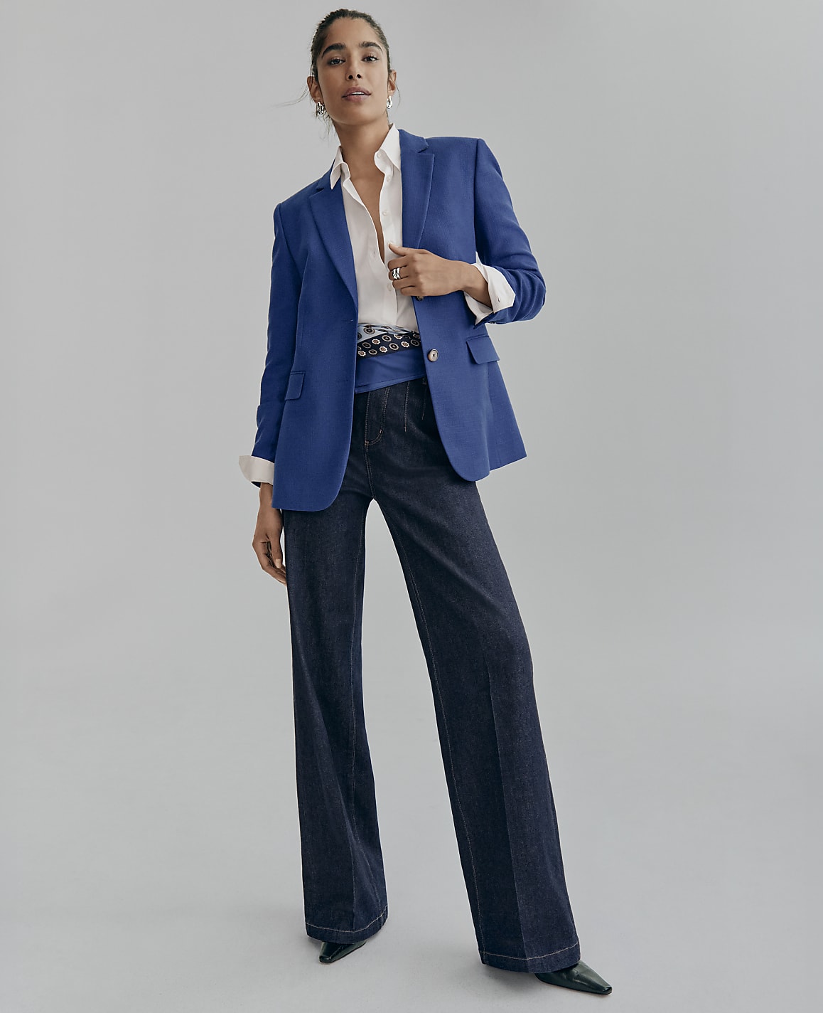 A woman wearing a blue blazer, white shirt, and wide-leg dark denim jeans stands against a plain light gray background.