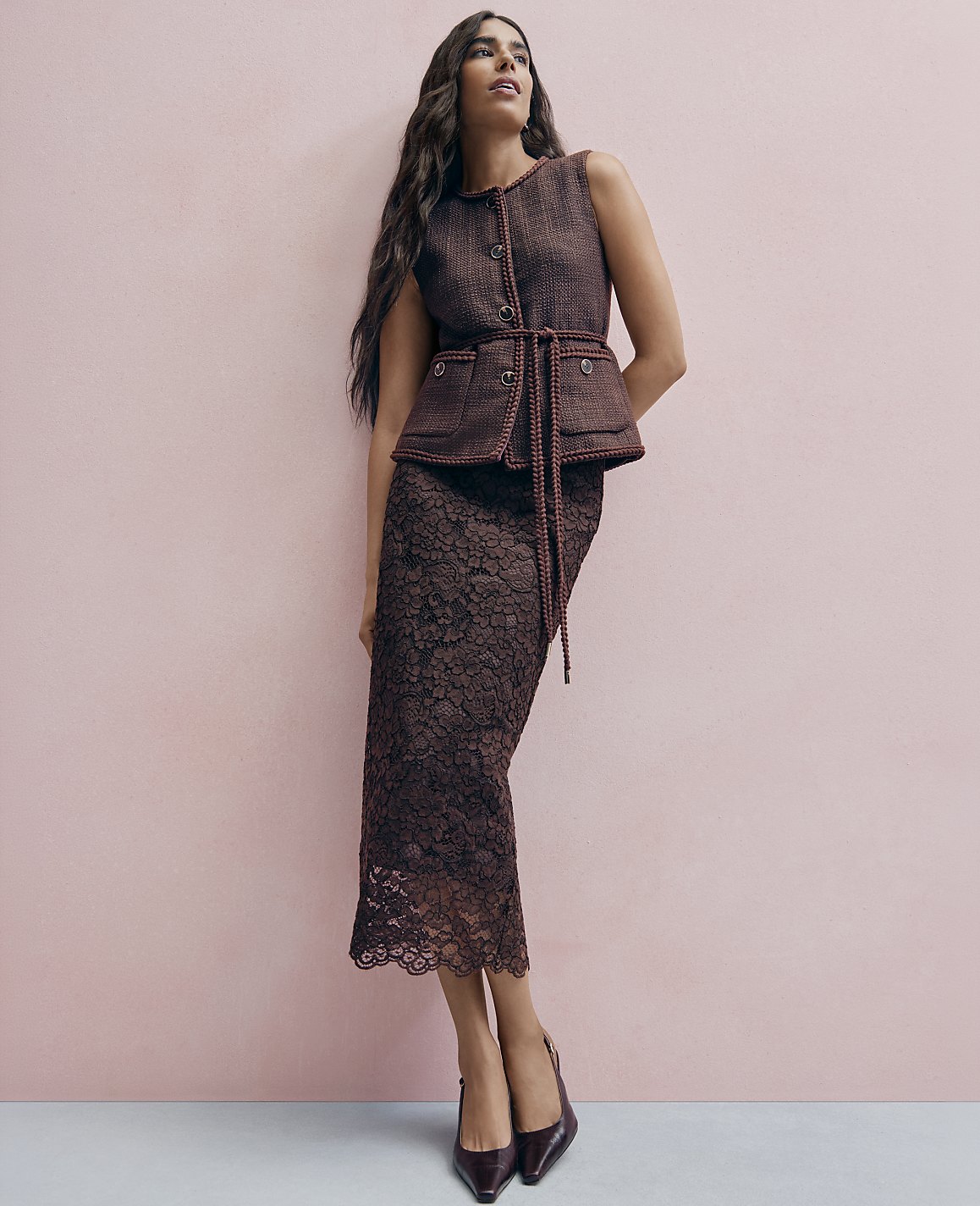 A woman wearing a brown tweed vest and lace skirt stands against a solid pink wall.