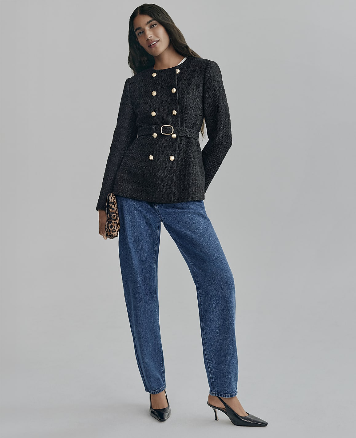 A woman wearing a belted black double-breasted jacket and blue jeans stands against a plain grey background.