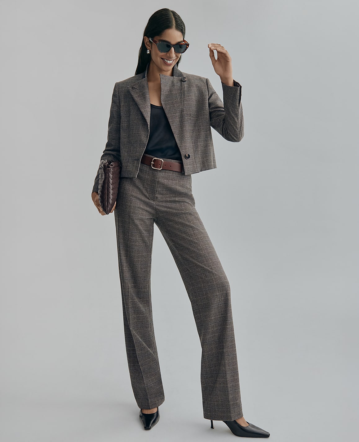 A woman wearing a grey plaid cropped blazer and matching trousers stands against a plain light grey background.