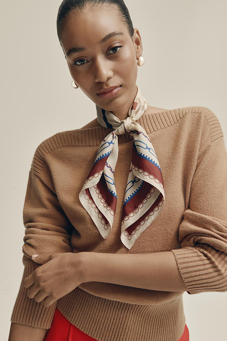 A woman wearing a tan sweater and a patterned silk scarf poses against a plain, light-colored background.