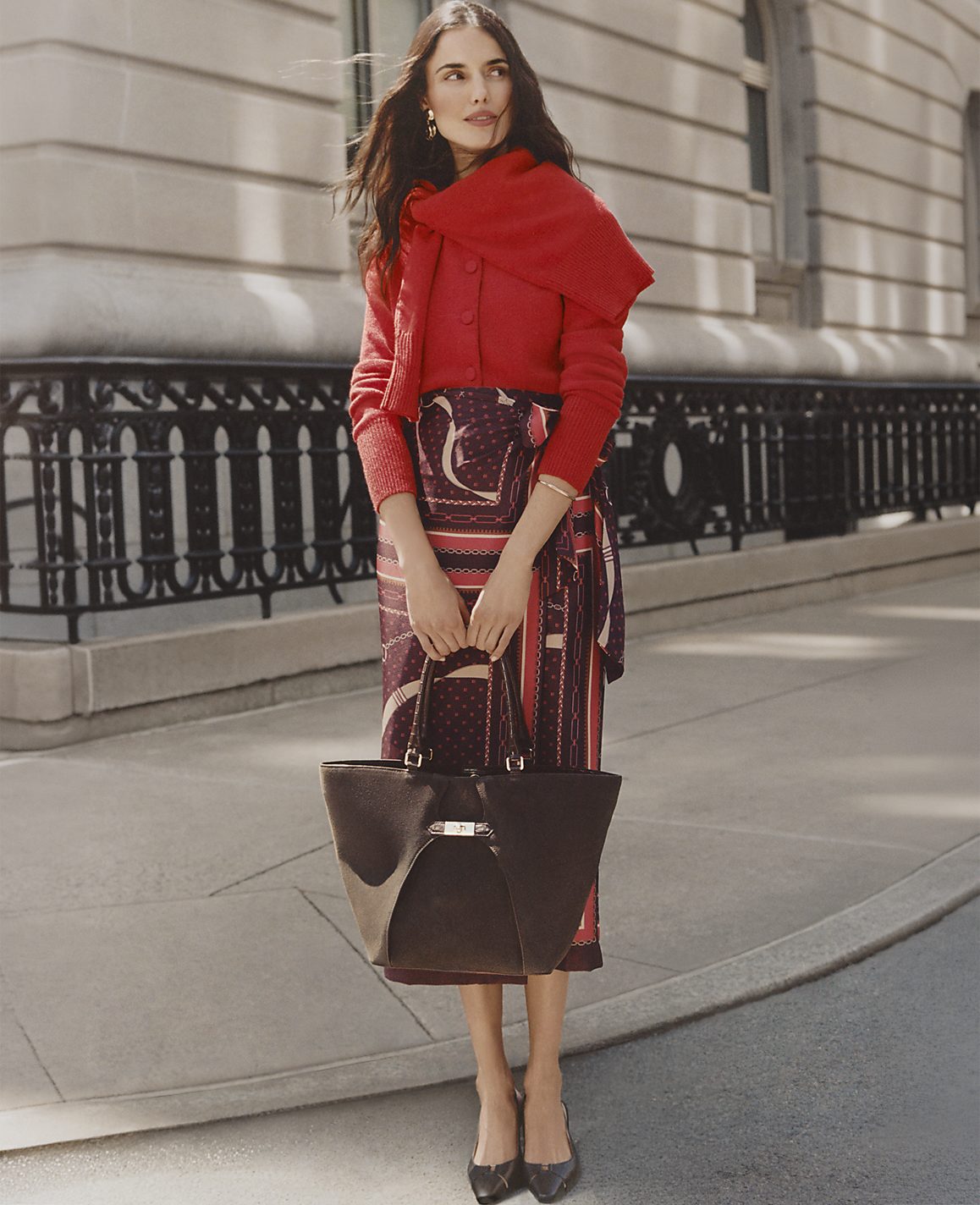 A woman wearing a red cardigan and a patterned skirt stands on a city sidewalk in front of a stone building with a black wrought-iron fence.