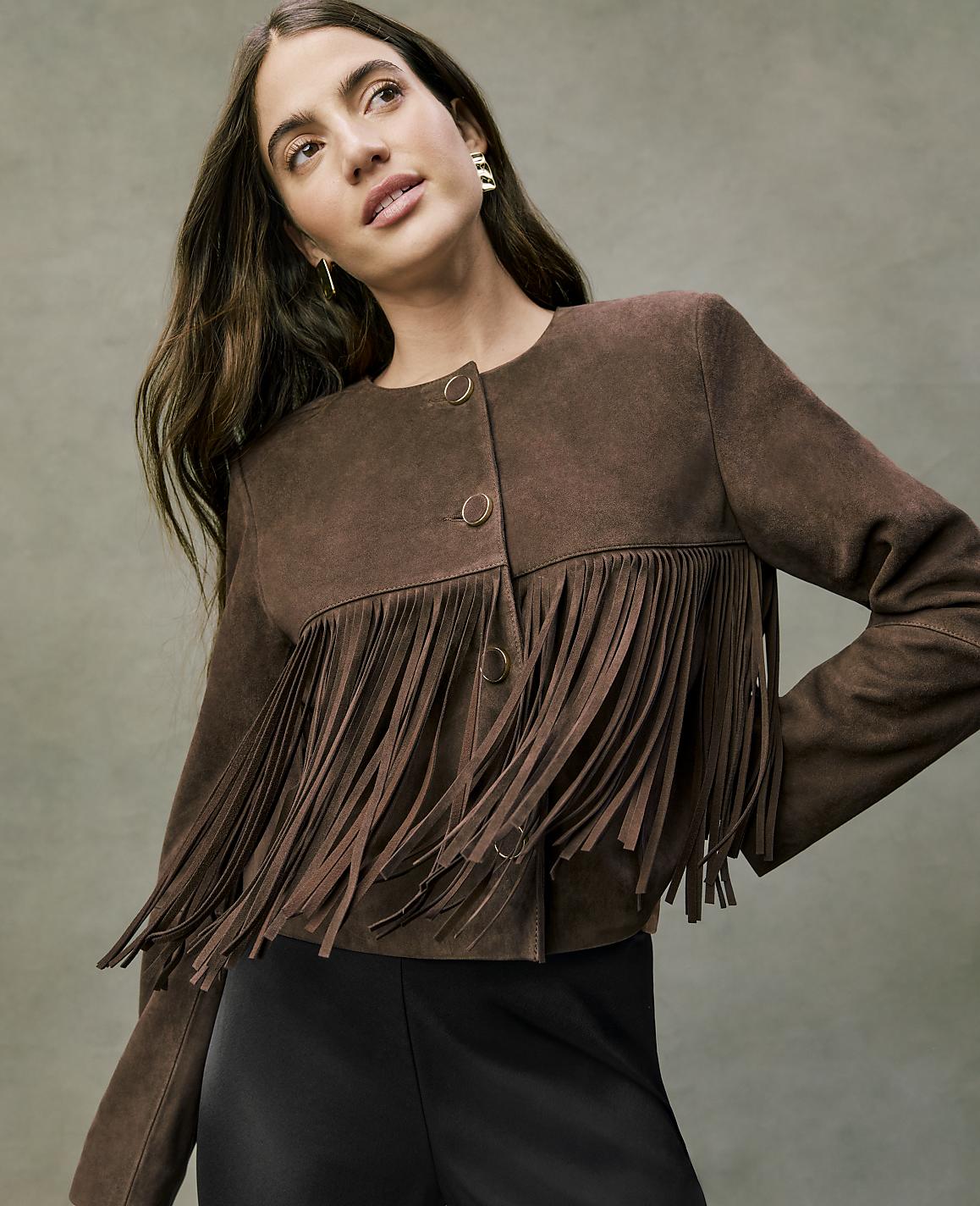 A woman wearing a brown suede fringe jacket and black pants poses against a neutral, textured grey background.