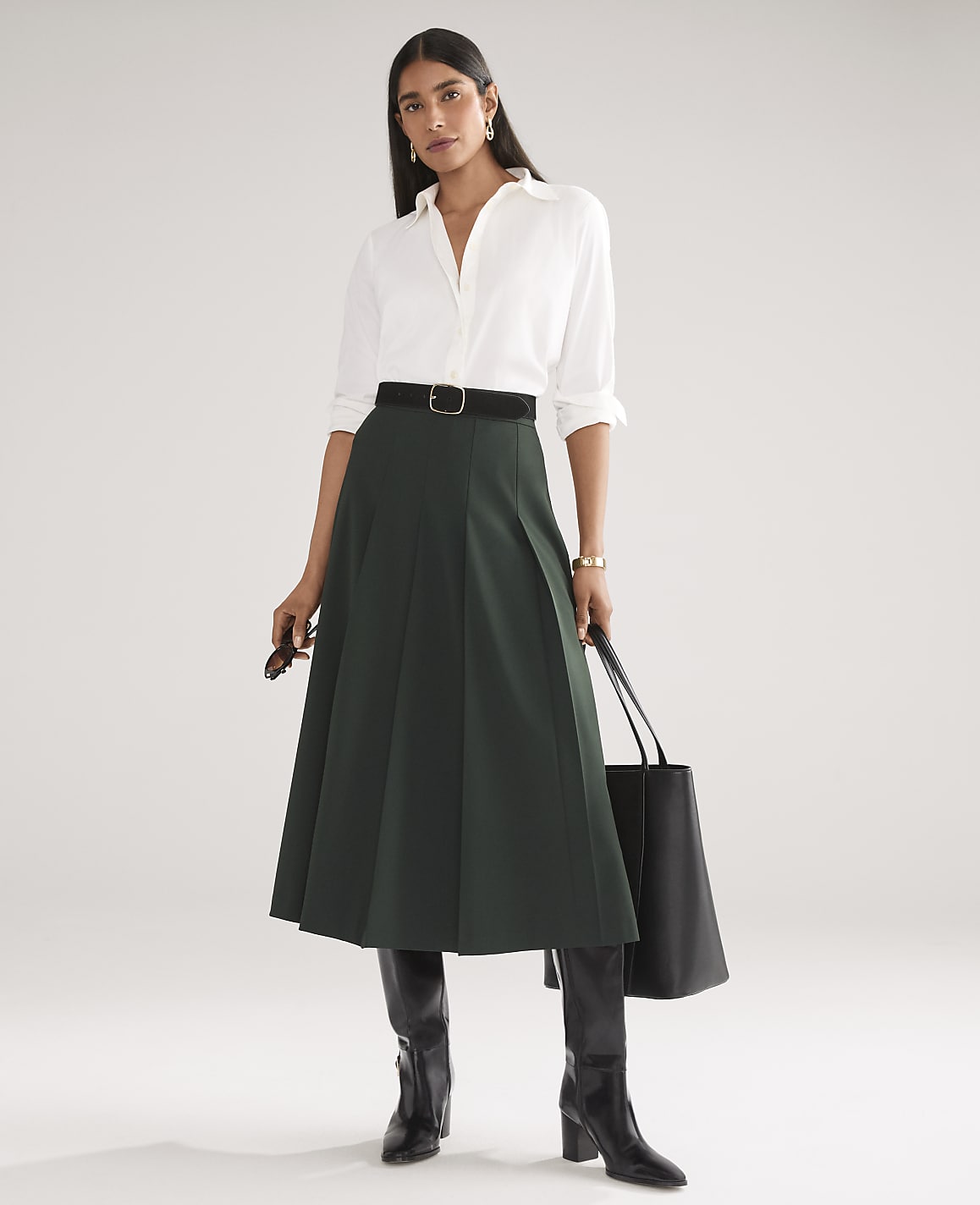 A woman wearing a white button-down shirt, a dark green pleated midi skirt, and black boots stands against a plain white background.