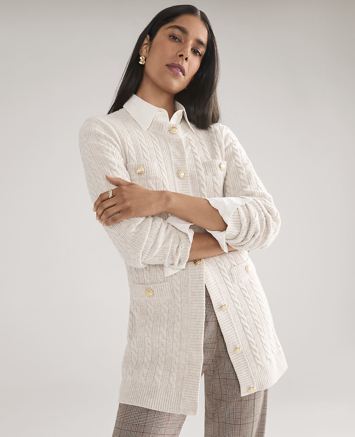 A woman wearing a white cable-knit cardigan over a collared shirt and plaid trousers poses against a plain light gray background.