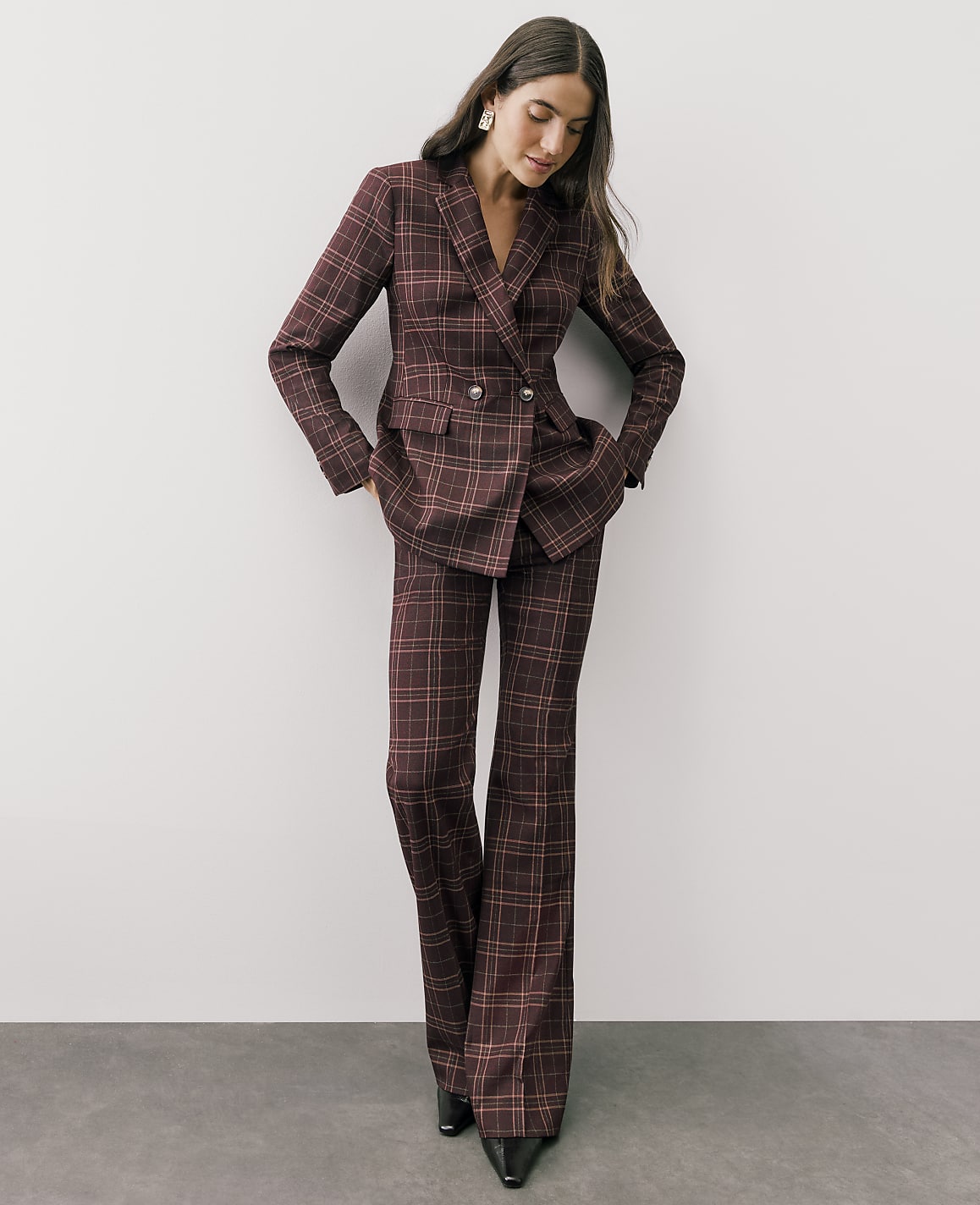 A woman wearing a burgundy plaid pantsuit stands against a plain white wall and grey floor.