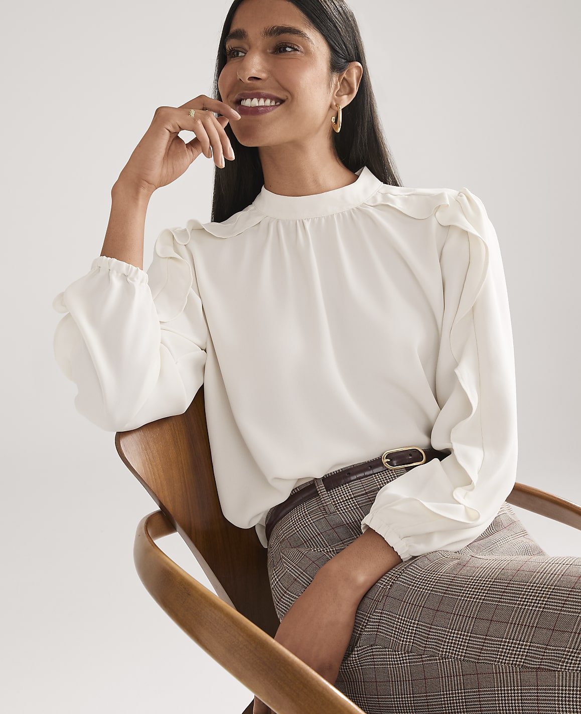 A smiling woman wearing a white ruffled blouse and plaid trousers sits in a wooden chair against a plain white background.