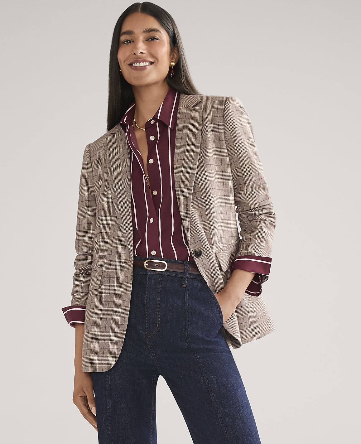 A smiling woman wearing a plaid blazer, striped maroon shirt, and dark blue jeans stands against a plain light gray background.