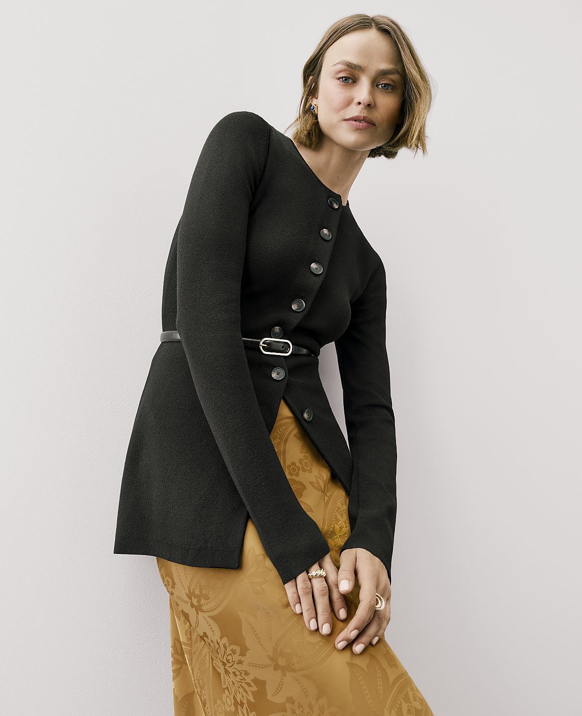 A woman wearing a black button-down jacket with a belt and a gold patterned skirt stands against a plain light gray background.