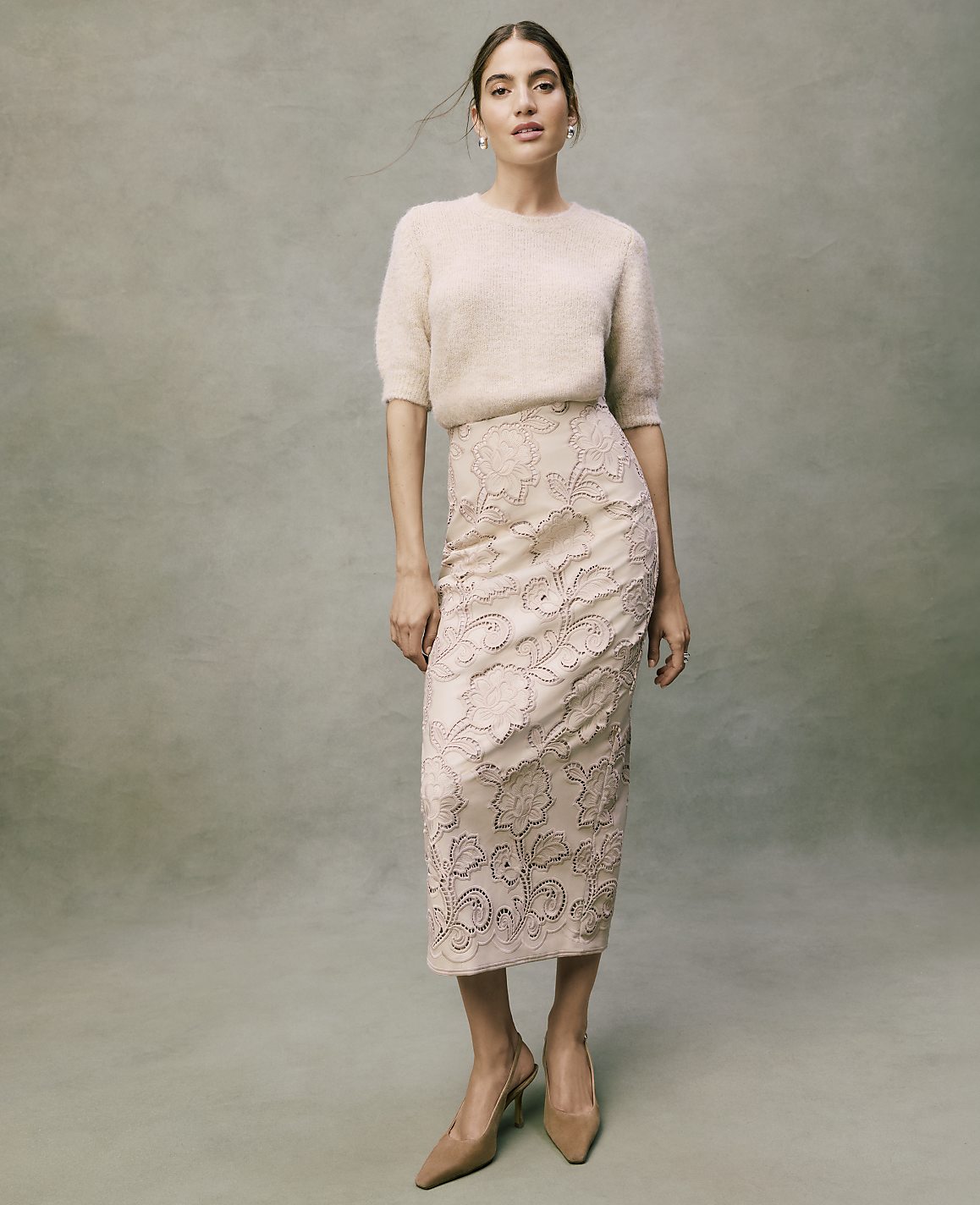 A woman wearing a fuzzy cream sweater, an embroidered pale pink skirt, and nude heels stands against a textured grey background.