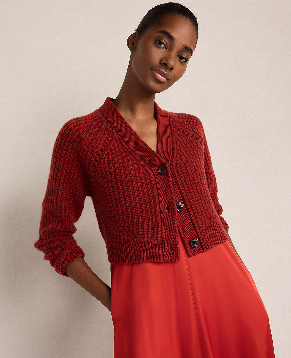 A woman wearing a red knit cardigan and a matching red satin skirt poses against a plain, neutral-toned background.