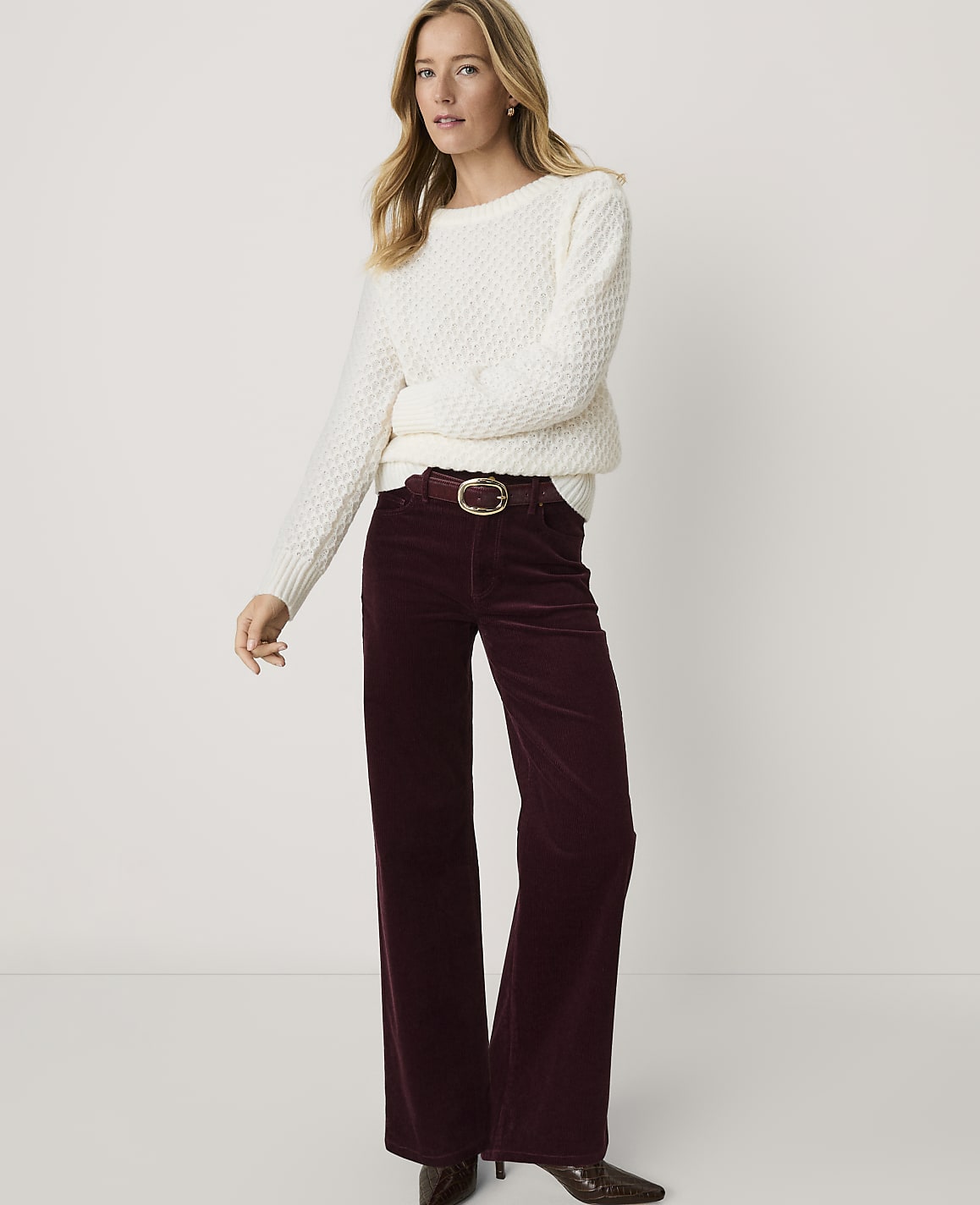 A woman wearing a white textured sweater and wide-leg burgundy corduroy pants stands against a plain white background.